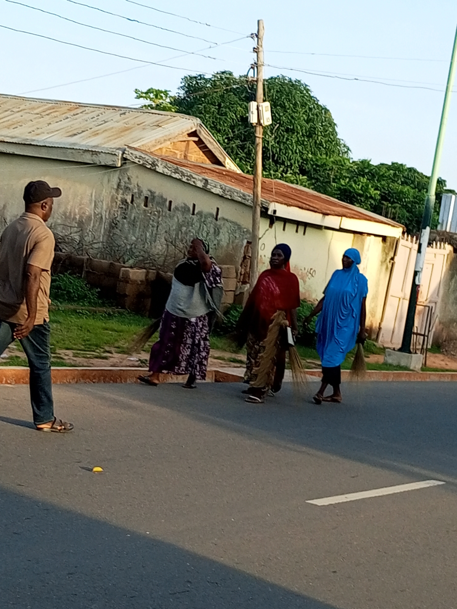 In Gombe, Nigeria, a man wearing a brown t-shirt and blue jeans, with a black baseball cap, stands on the left side of a paved road. He is facing away from the viewer, towards three women. To his right, across the road, are the three women. The first woman is wearing a dark headscarf and a patterned purple dress, holding a broom. The second woman is dressed in red with a head covering and is also holding a broom. The third woman, on the far right, is wearing a bright blue headscarf and a blue dress, and is also carrying a broom. They appear to be walking or standing in the street. Behind them is a building with a corrugated metal roof and a light-colored wall. Trees and a utility pole are also visible in the background. The scene takes place during the day, as indicated by the lighting and shadows.