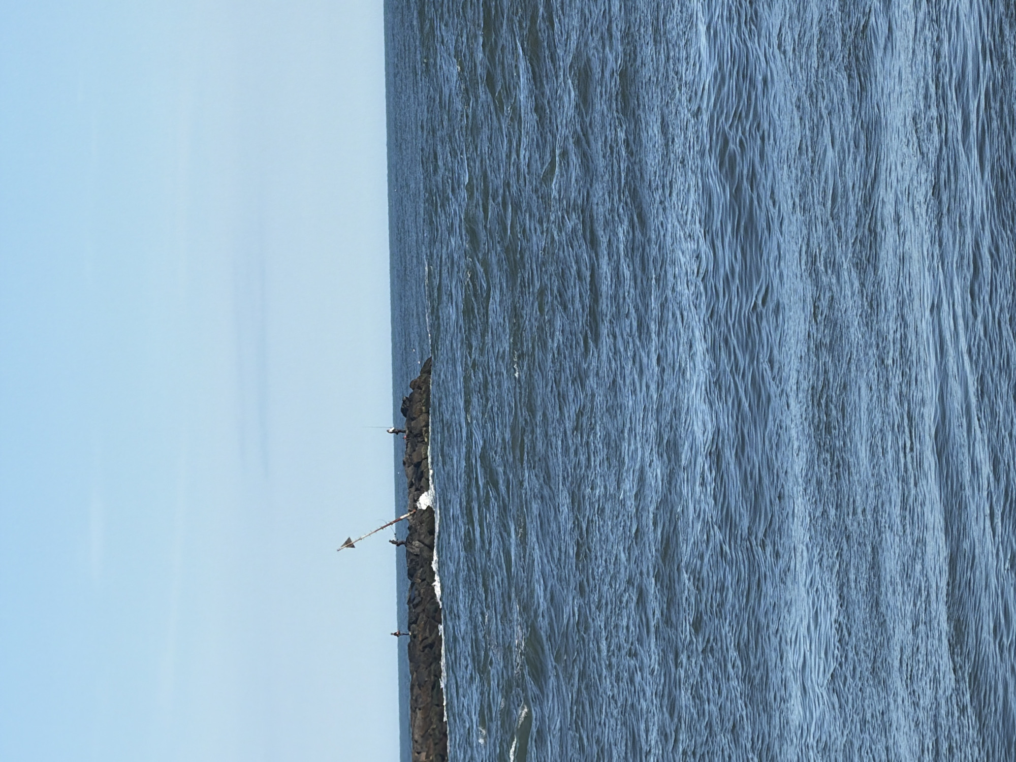 The image shows a rocky jetty extending into the ocean under a clear blue sky. Several individuals are fishing on the jetty. The water is choppy with visible waves. A partially broken navigational marker with a white triangular sign is leaning precariously on the jetty. The scene is located in Hampton Bays, New York.