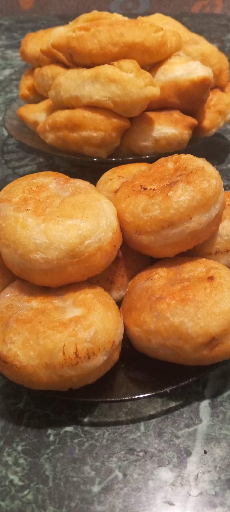 A close-up, overhead shot shows a pile of golden-brown fried dough pastries on a dark green, speckled countertop. The pastries are arranged on two dark plates, with a stack of elongated, pillow-like pastries on the upper plate and several round, flatter pastries in the foreground. The lighting is dim, casting subtle shadows and highlighting the oily sheen on the pastries. The background is out of focus, but the countertop appears to be marble or a similar material. There are no people or any indication of a specific location within the city of Dyurtyuli, Russia.