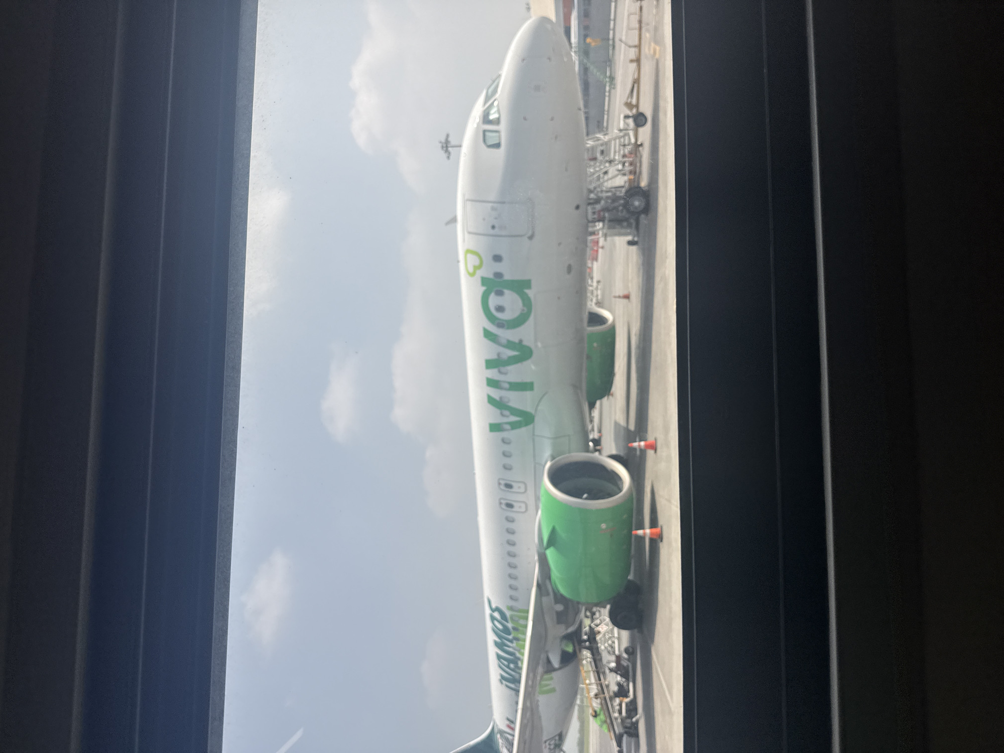 The image shows a Viva Aerobus airplane viewed through a window. The plane is white with green accents, parked on an airport tarmac. In the background, are other airport structures and a cloudy sky. The photo was likely taken in Apodaca, Mexico.