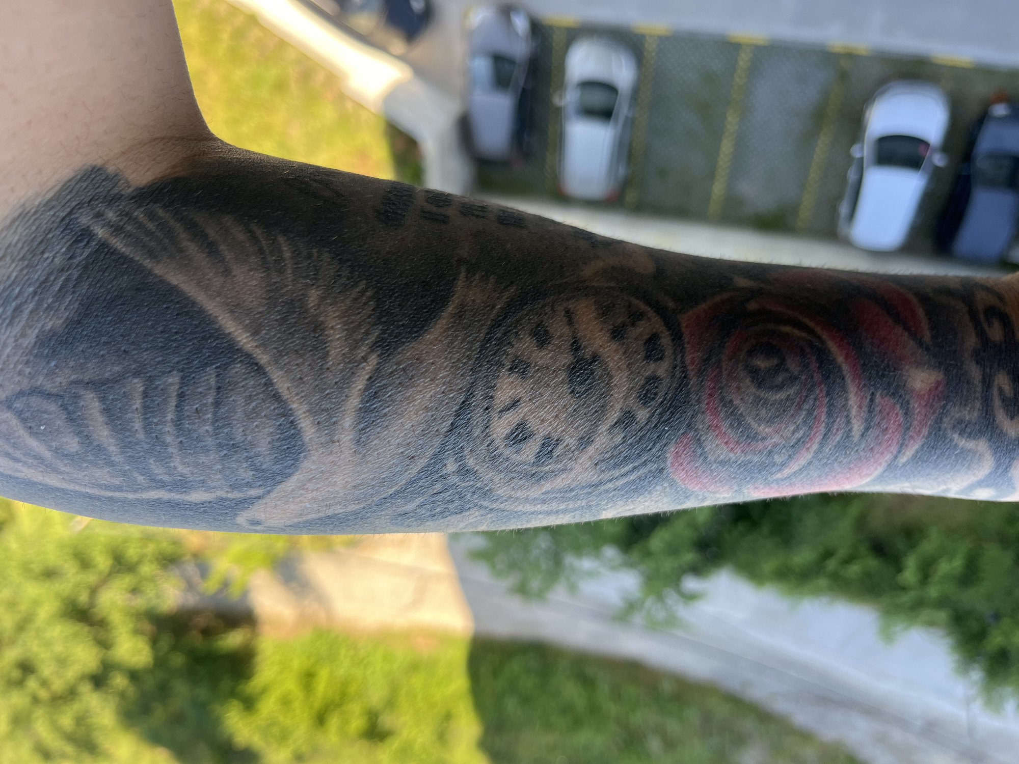 The image shows a person's tattooed forearm with a view of what appears to be an urban environment. The tattoos include what looks like a rose and a clock. In the background, there is a parking lot with several cars, and trees, suggesting the image was taken from a high vantage point, possibly a balcony. It is not possible to identify this as Varna, Bulgaria based on the image provided.