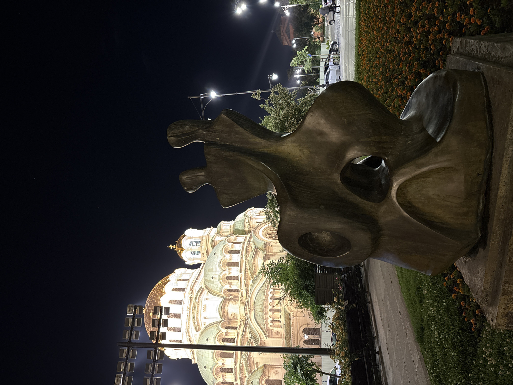 This nighttime image captures a dramatic scene in Sofia, Bulgaria, featuring the illuminated St. Alexander Nevsky Cathedral and a prominent abstract sculpture.
The main subject on the left is the magnificent St. Alexander Nevsky Cathedral, bathed in warm, artificial light that highlights its intricate architectural details. Multiple domes, some gleaming gold and others a pale greenish-blue, rise above a complex structure of arches, windows, and decorative elements. The cathedral's grandeur stands out against the dark night sky.
In the foreground, occupying the right side of the frame, is a large, bronze-colored abstract sculpture. Its smooth, undulating surfaces and organic, flowing shapes create a sense of movement, with several open spaces or voids within its form. The sculpture's modern aesthetic contrasts with the historical architecture of the cathedral.
Between the sculpture and the cathedral, some trees and green foliage are visible, partially illuminated. To the right of the sculpture, a paved pathway or sidewalk leads towards the background. Alongside it, a bed of vibrant orange flowers, possibly marigolds, adds a burst of color. Further back on the right, bright streetlights cast their glow on a street or plaza, revealing outlines of parked vehicles and distant buildings.
The time of day is clearly night, with the sky being a deep, dark blue-black. The scene is lit entirely by artificial sources: the floodlights on the cathedral, the streetlights, and subtle ambient light elsewhere. There is no visible activity or specific event taking place, and no text is discernible in the image. The overall impression is one of architectural beauty and artistic contrast under the nocturnal sky.