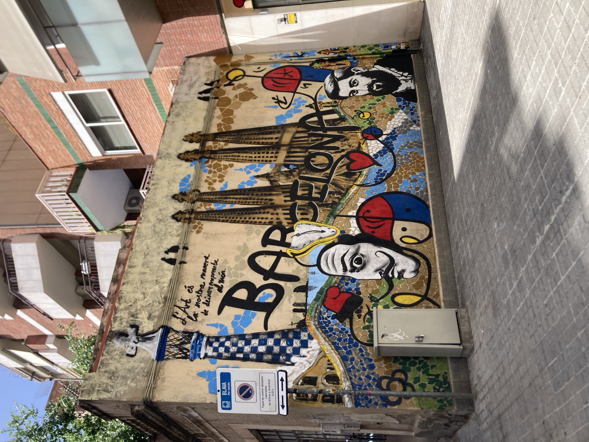 The image shows a colorful mural painted on the exterior wall of a building on a street in Barcelona, Spain. The mural depicts iconic Barcelona landmarks and figures. On the left, a representation of the Sagrada Familia's spires is visible, and the word "BARCELONA" is prominently displayed in large, bold letters. To the left of the Sagrada Familia depiction, there's a lighthouse-like structure with a checkered pattern. A portrait of Salvador Dalí, with his signature mustache and melting clock, is a central feature of the mural. Another portrait, believed to be of Antoni Gaudí, is on the right side. The lower part of the mural is a mosaic of colorful tiles, forming a wavy pattern and various circular designs. There are also stylized depictions of birds and hearts. A street sign with Catalan text, indicating parking restrictions, is mounted on a pole in front of the mural. A grey electrical box is situated at the base of the wall, partially obscuring the mural. The street itself is paved with grey rectangular tiles. Buildings with balconies are visible in the background, suggesting an urban setting. The lighting indicates it is daytime, with shadows cast on the street.