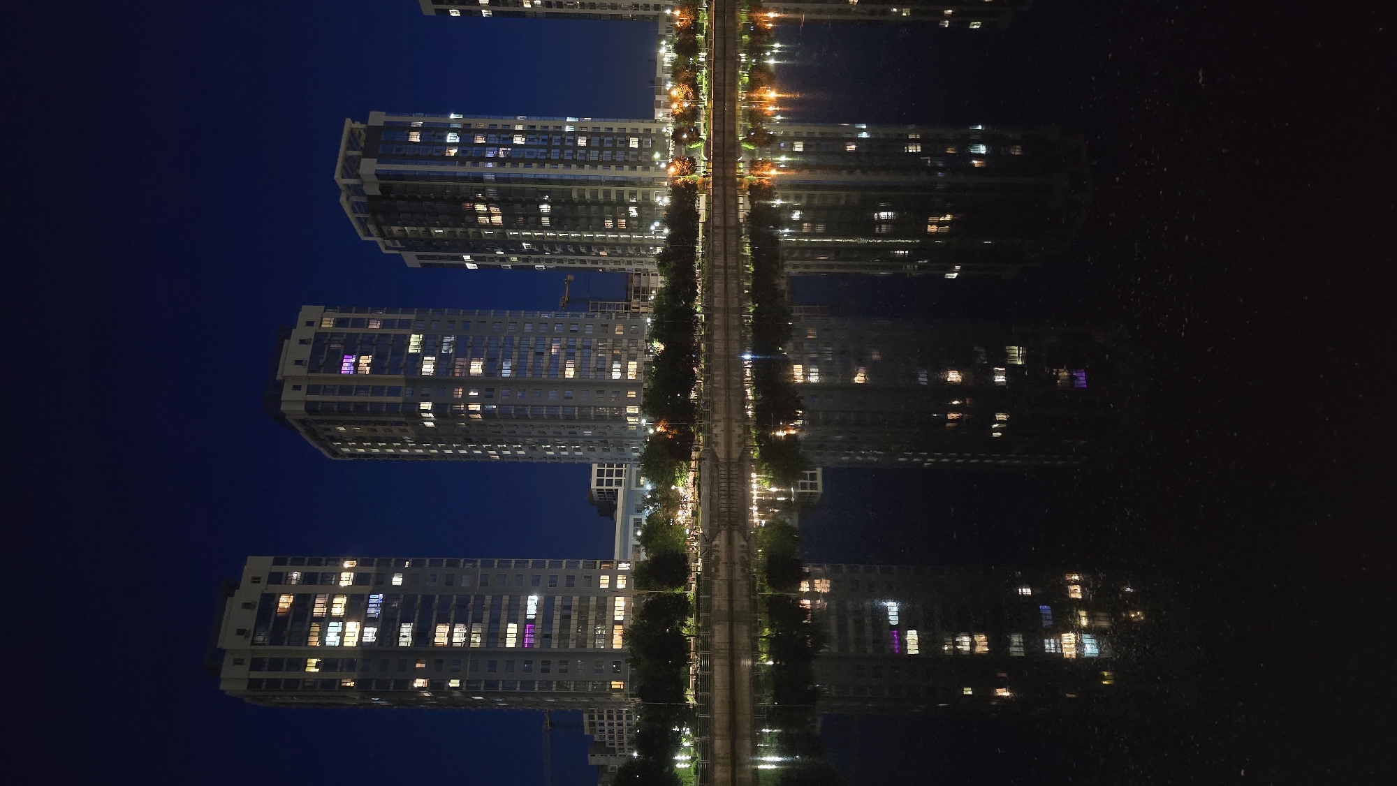 This image presents a tranquil night scene of a modern cityscape, characterized by its striking reflections in a calm body of water. The perspective is oriented with the city on the left and its reflection on the right, separated by a central linear feature.
On the left, multiple tall, contemporary residential buildings or apartment blocks rise against a deep indigo-blue night sky. Numerous windows across these structures are illuminated, displaying a mix of warm yellow, bright white, and occasionally purple or blue lights, suggesting active habitation within the multi-story buildings.
Between the buildings and the water, a well-lit promenade or road stretches horizontally, adorned with trees and bright streetlights that cast warm, yellowish glows. This central strip forms a clear division between the real structures and their mirrored counterparts.
To the right, a still body of water acts as a perfect mirror, reflecting the entire cityscape with remarkable clarity. The inverted images of the illuminated windows, streetlights, and the general outline of the buildings are sharply rendered on the water's surface, creating an almost symmetrical composition. Subtle textures in the reflection indicate the water's surface, particularly towards the right edge.
The scene is set at night, with the sky exhibiting a dark blue hue rather than pure black, suggesting either late dusk or early night. The weather appears clear and calm, allowing for the pristine reflections. While no direct human activity is visible, the multitude of lit windows conveys a sense of a bustling urban environment under a peaceful night sky. No discernible text is present in the image. The setting is a modern urban waterfront, consistent with areas in cities like Kyiv, Ukraine.