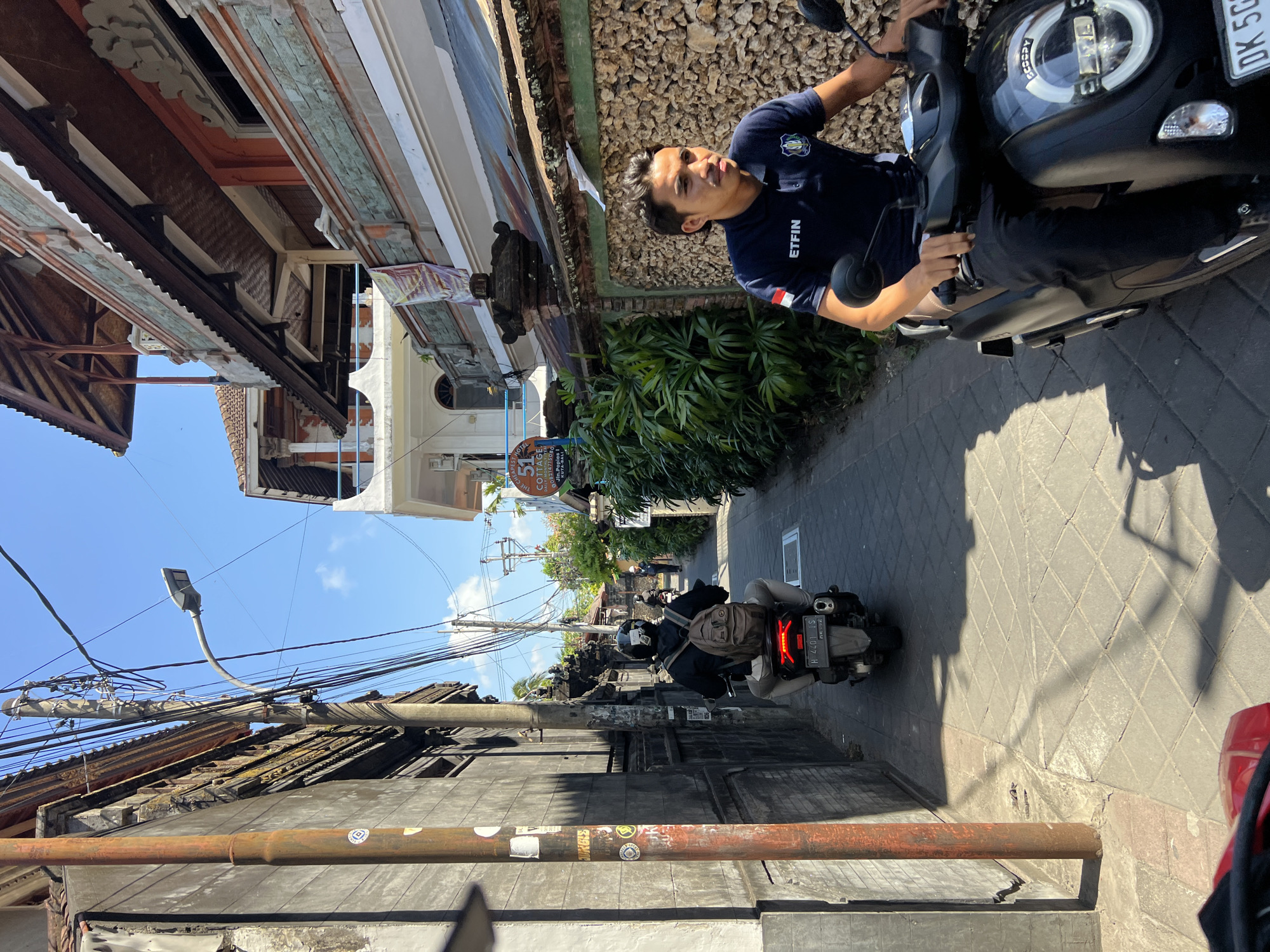 A narrow alleyway in Kuta, Indonesia, is depicted on a sunny day. Two men are riding scooters. In the foreground, a man in a navy blue polo shirt with "ETFIN" printed on it rides a black scooter. Behind him, another man wearing a backpack rides a scooter. Buildings line both sides of the alley. One building has a sign that says "51 COTTAGE". The sky is clear and blue.