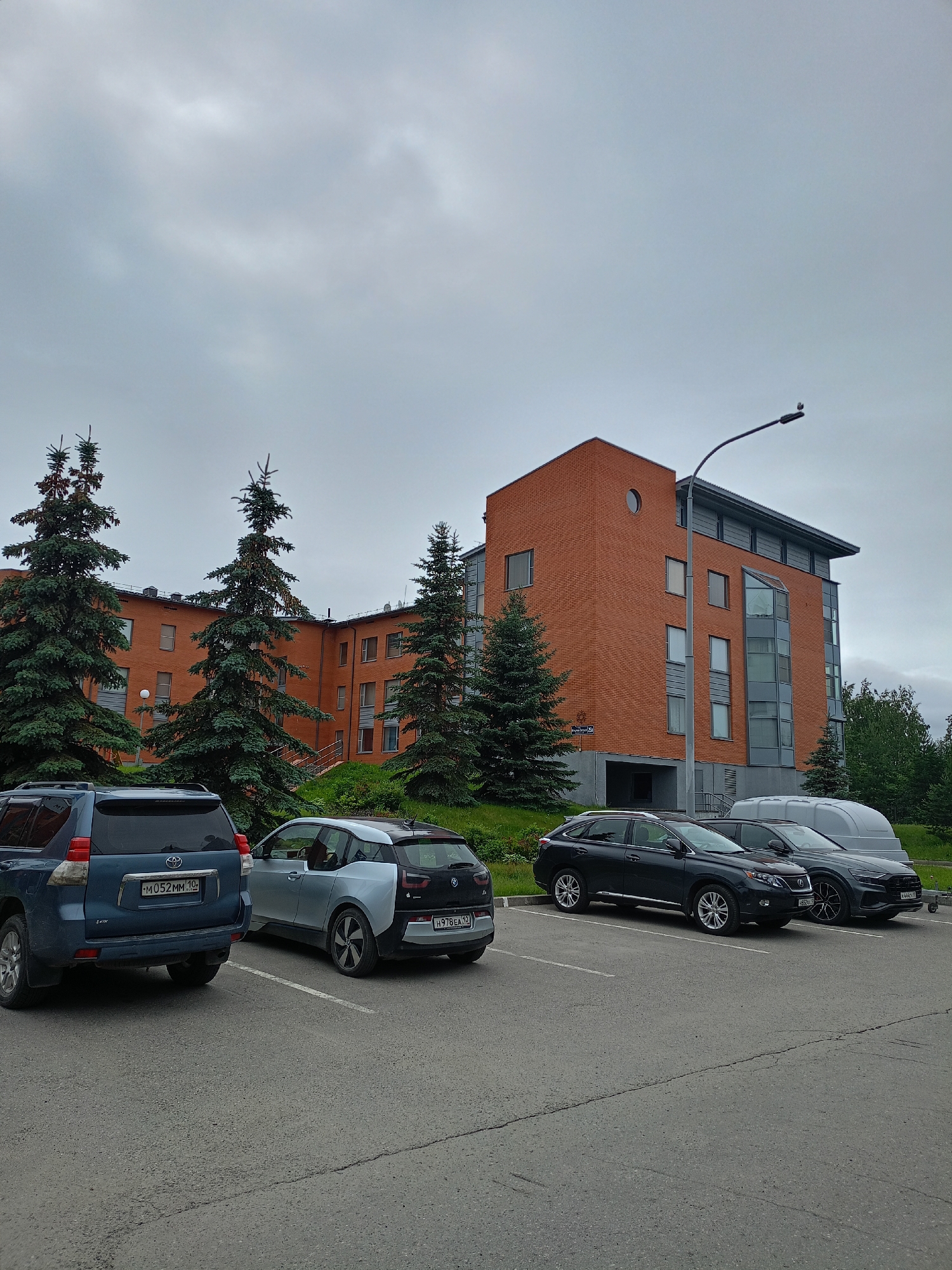The image shows a parking lot in front of a modern brick building. The building has multiple stories with windows and a distinctive circular window on one of the upper floors. Several evergreen trees are planted in front of the building. In the parking lot, there are several cars.
The cars visible include a blue SUV, a silver and black hatchback, a dark-colored SUV, and a dark-colored sedan. Two of the cars have visible license plates. The blue SUV's license plate reads "M052MM 10". The silver and black hatchback's license plate reads "H978EA 32". Another car's license plate reads "V 4440C7 150".
A street sign is visible on the building, with text that appears to be in Cyrillic. A lamppost stands near the building, extending into the sky. The sky is overcast, suggesting cloudy or possibly rainy weather. The overall scene appears to be a typical day in a residential or commercial area. No people are visible in the image. The time of day is indeterminate due to the overcast sky.