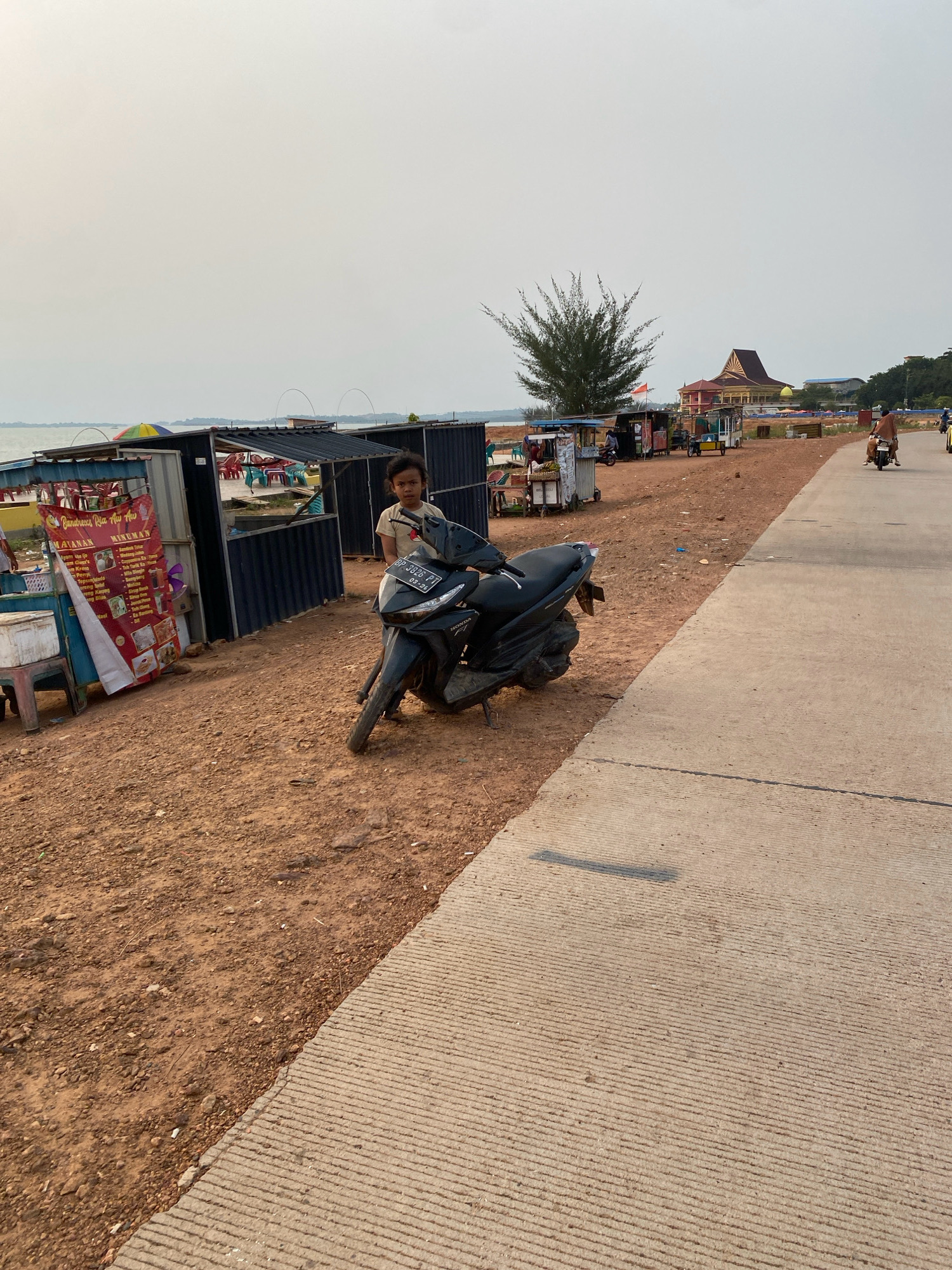 A young boy with curly hair stands next to a black motorcycle in front of a row of food stalls by the water in Tanjung Pinang, Indonesia. The stalls have metal roofs and are decorated with colorful umbrellas and signs. The ground is a mix of dirt and gravel, and a paved walkway runs along the right side of the image. In the background, there is a large building with a distinctive roof, a pine tree, and other structures. Several people are seen on motorcycles in the distance. The sky is overcast, and the overall atmosphere appears to be hazy.