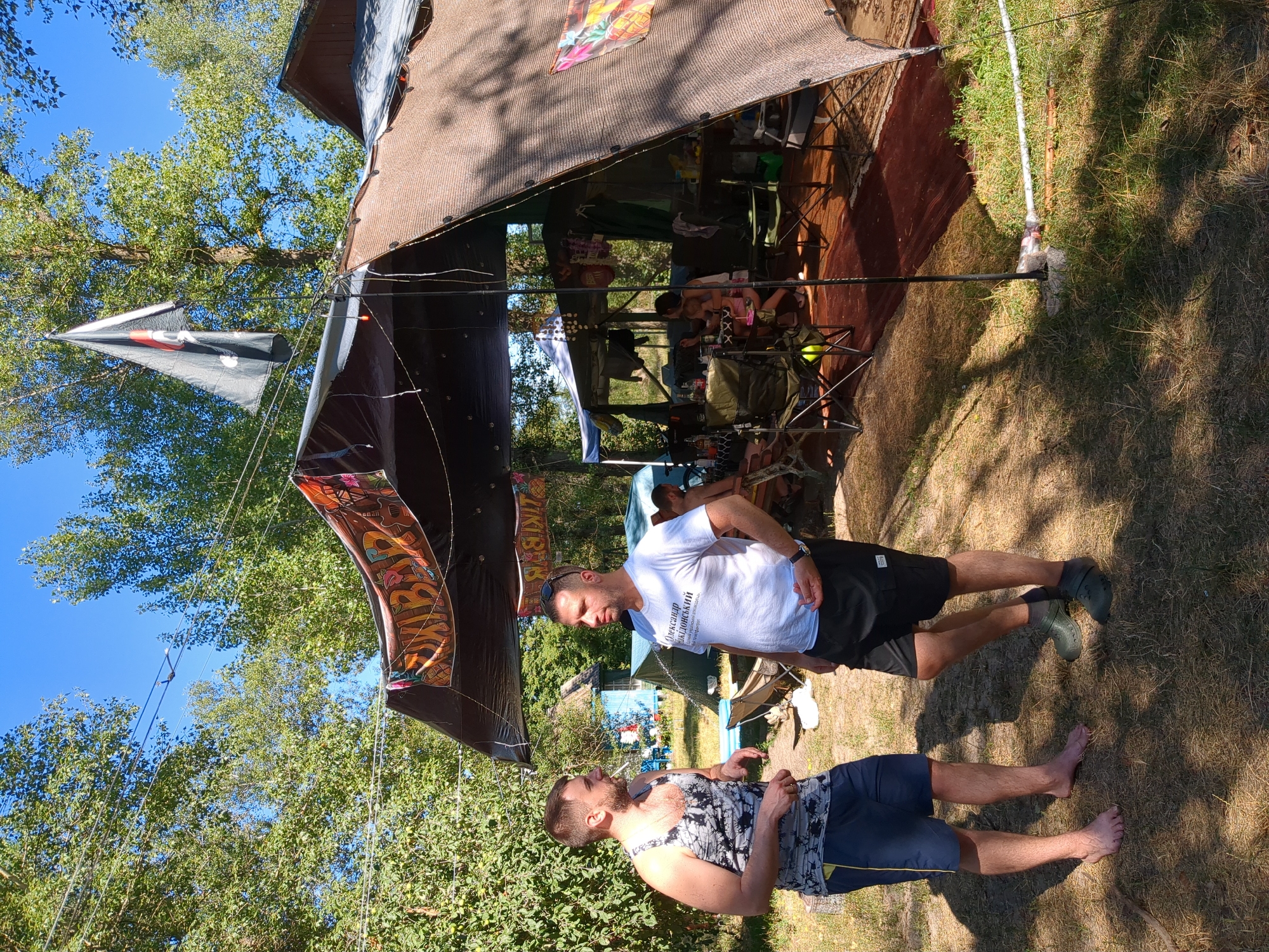 Two men are standing in a grassy area near a large, open-sided tent with a dark brown mesh-like covering. The man on the left wears a gray and white patterned tank top and navy blue shorts, and is barefoot. The man on the right wears a white t-shirt with Ukrainian text and black shorts, and green clogs. Behind them, more tents and trees are visible. The tent has a banner with colorful graphics and text, and a pirate flag is flying above it. Inside the tent, several people are seated, and there are tables and chairs. The scene appears to be an outdoor gathering or festival.