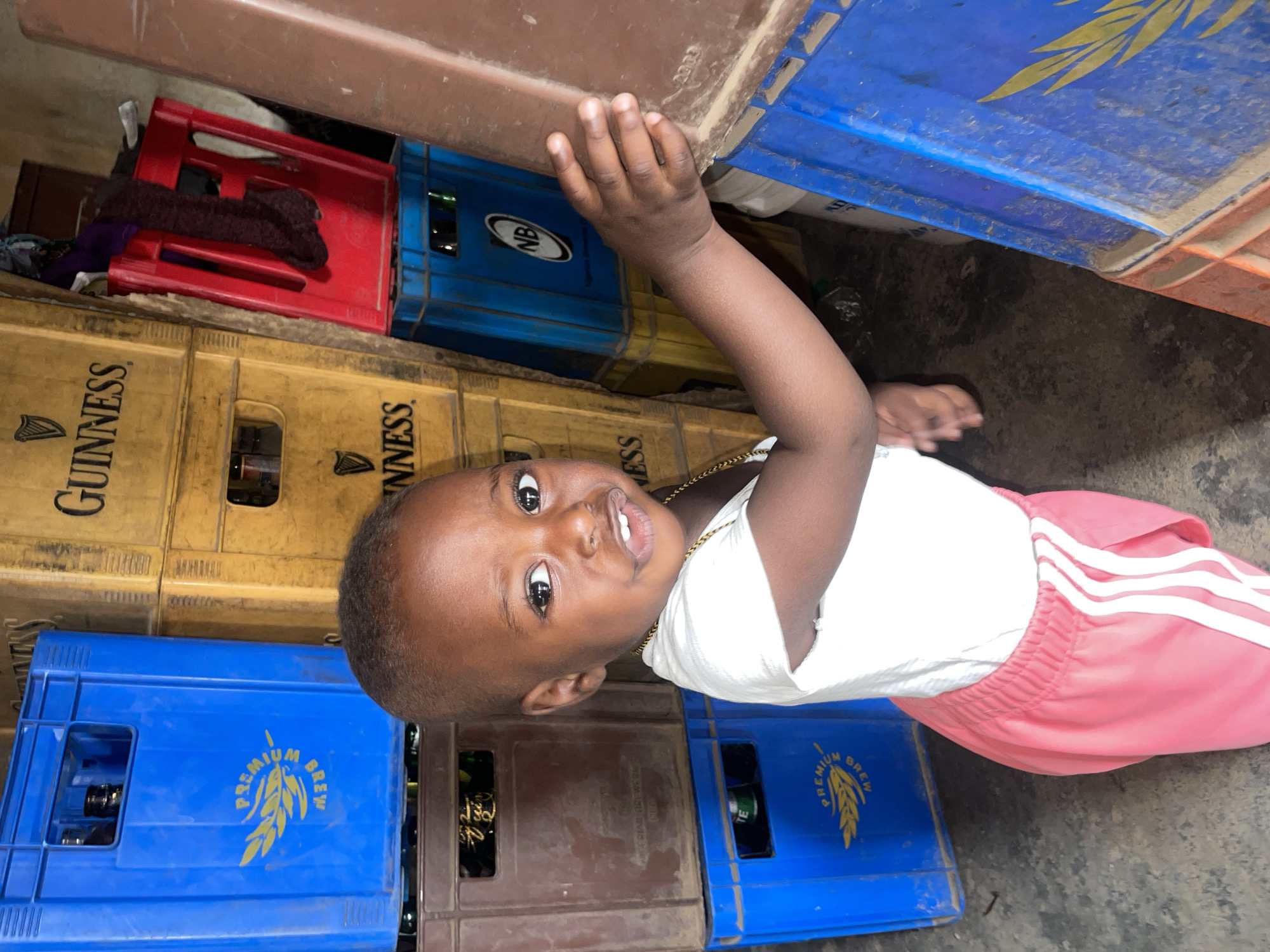 This image, taken in Jos, Nigeria, features a young child standing amidst a collection of beverage crates. The child, with dark skin, dark hair, and bright eyes, wears a white t-shirt, a light-colored necklace, and red/pink shorts with white stripes. They are looking directly at the camera with a neutral to slightly smiling expression. Their left hand is raised, grasping the underside of a brown, flat surface positioned above their head.
The setting appears to be an indoor or semi-enclosed space, possibly a storage area or a small shop. The floor is a dark, rough surface. Numerous plastic beverage crates are stacked around the child. Visible brands and texts on the crates include "GUINNESS" with its harp logo on yellow/beige crates, and "PREMIUM BREW" with a wheat motif on blue crates. Some blue crates also show the "NB" logo, indicating Nigerian Breweries, which is consistent with the Jos, Nigeria context. A red crate, containing what looks like folded dark fabric, is also present in the upper left. The year "2003" is faintly visible on one of the upper crates. The overall lighting suggests an indoor environment.