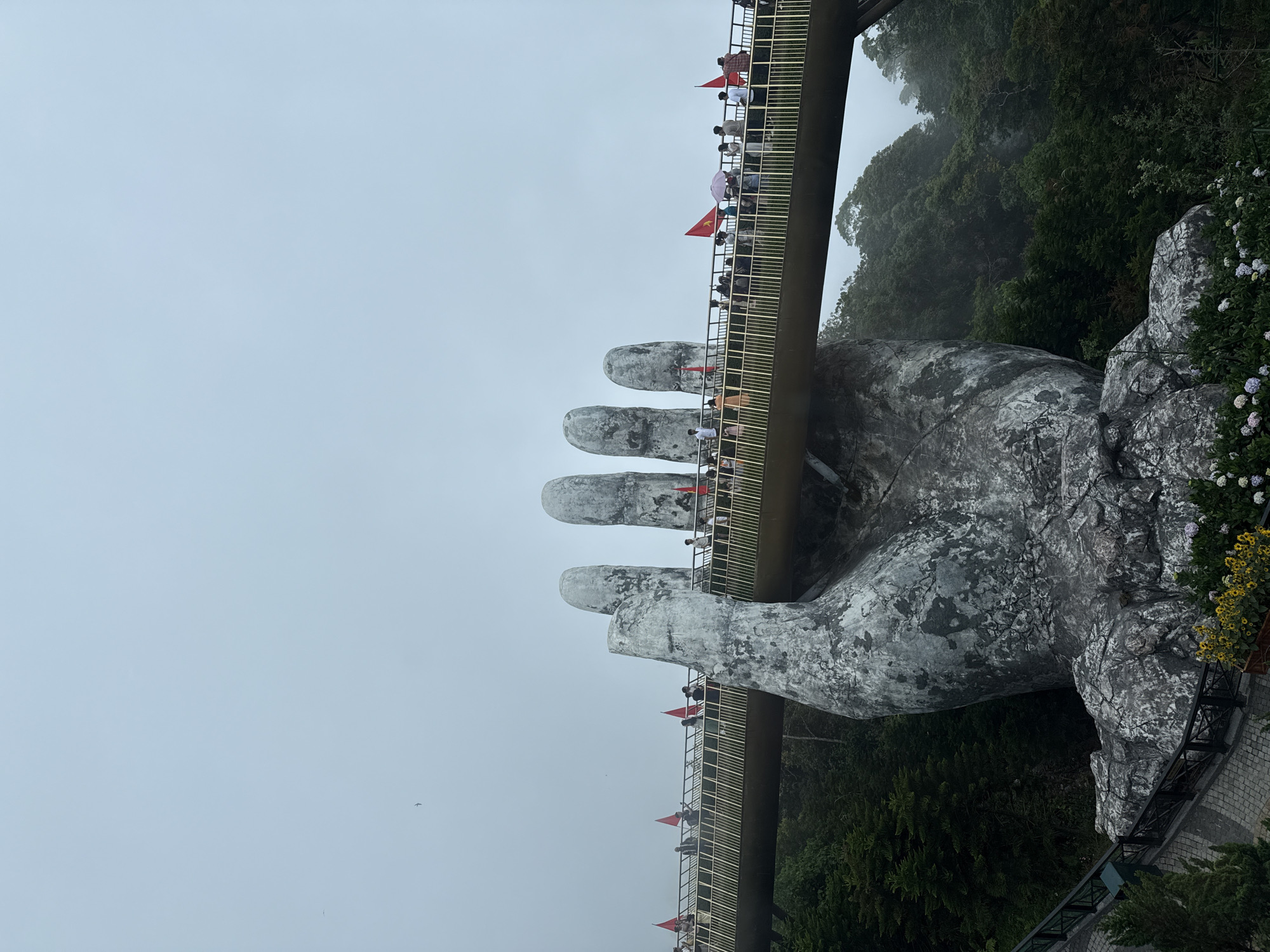 This photograph captures the iconic Golden Bridge (Cầu Vàng) in Da Nang, Vietnam, an architectural marvel appearing to be held aloft by two colossal, weathered stone hands. The bridge, with its distinctive golden railing, spans across the upper-mid section of the frame. Numerous individuals are visible walking along the bridge's pathway, suggesting a popular tourist attraction.
The scene is set on an overcast or foggy day, evidenced by the uniformly light grey-blue sky that fills a significant portion of the background, contributing to a muted and slightly hazy atmosphere. Below and to the right of the bridge, a lush, verdant mountain slope covered in dense trees and foliage descends into the misty distance. Large, textured rock formations are also visible as part of the natural landscape surrounding the structure. Several small red flags with a yellow star, identifiable as Vietnamese flags, are prominently displayed along the bridge's railings. The people on the bridge appear to be observing the views or simply enjoying a stroll. No discernible text is visible.