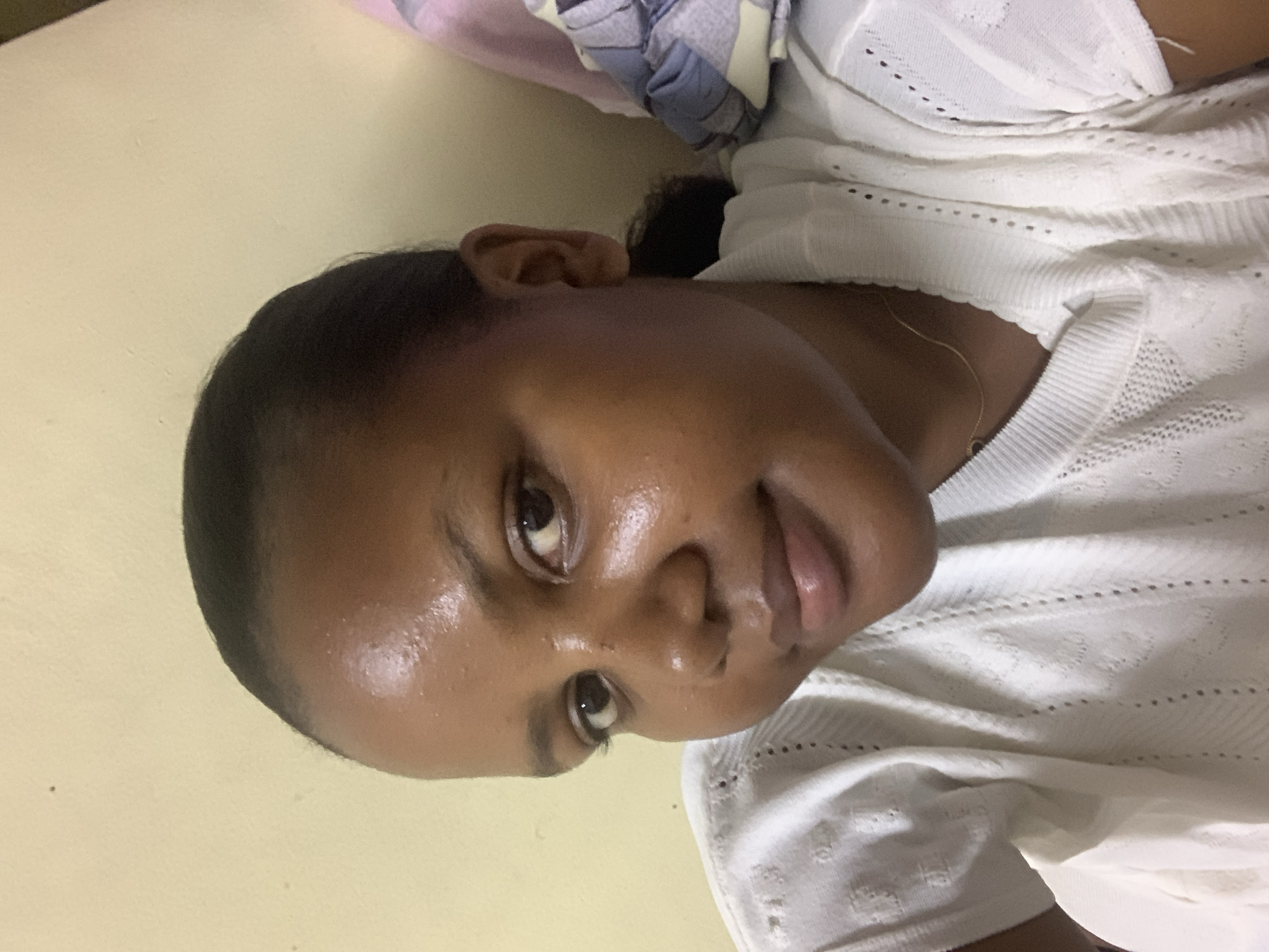 A close-up portrait of a woman with dark skin and short black hair. She has dark eyes and a slightly parted mouth. She is wearing a white crocheted top. The background is a plain light yellow wall. The location in Dar es-Salaam, Tanzania, is unknown.