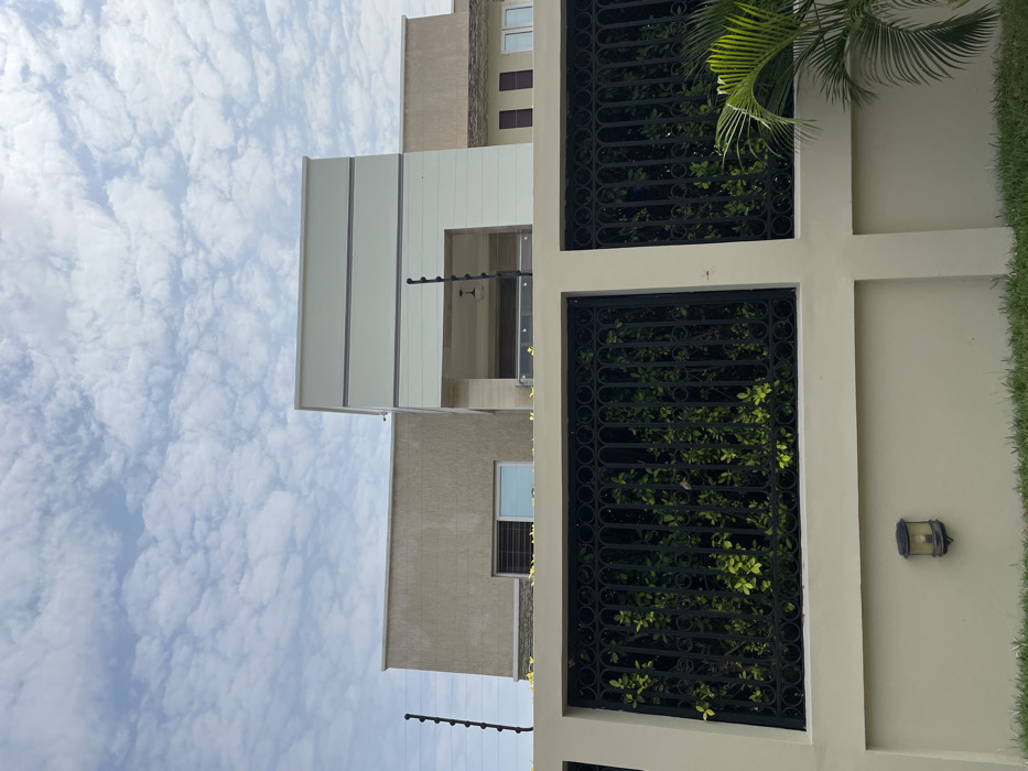 This image depicts the exterior of a modern, multi-story building, likely a residence, located in Adenta Municipality, Ghana, under a sky with scattered white clouds. The view is from a low-angle perspective, observing the structure from behind a perimeter wall.
In the foreground, a light beige concrete perimeter wall spans horizontally, topped with black electric fence posts and multiple strands of wire. Two rectangular sections of ornate black wrought iron fencing are integrated into the upper part of the wall, revealing dense green foliage immediately behind them. An amber-colored outdoor wall light fixture is affixed to the lower left portion of the concrete wall. A strip of green grass is visible at the base of the wall, and a partial view of a green palm frond is present on the far right.
The building behind the wall exhibits contemporary architecture with varied façade treatments. The left section of the building features a textured light brown/beige finish, with one horizontal rectangular window and one taller rectangular window visible. The central portion of the building presents a prominent off-white/light gray façade, accented with two dark gray horizontal stripes near its top. This central section includes a recessed area on the first floor, featuring a dark pendant light fixture and a clear glass railing. The right side of the building shows lighter-colored walls and several windows with reddish-brown frames. No individuals, explicit actions, or interactions are present in the scene.