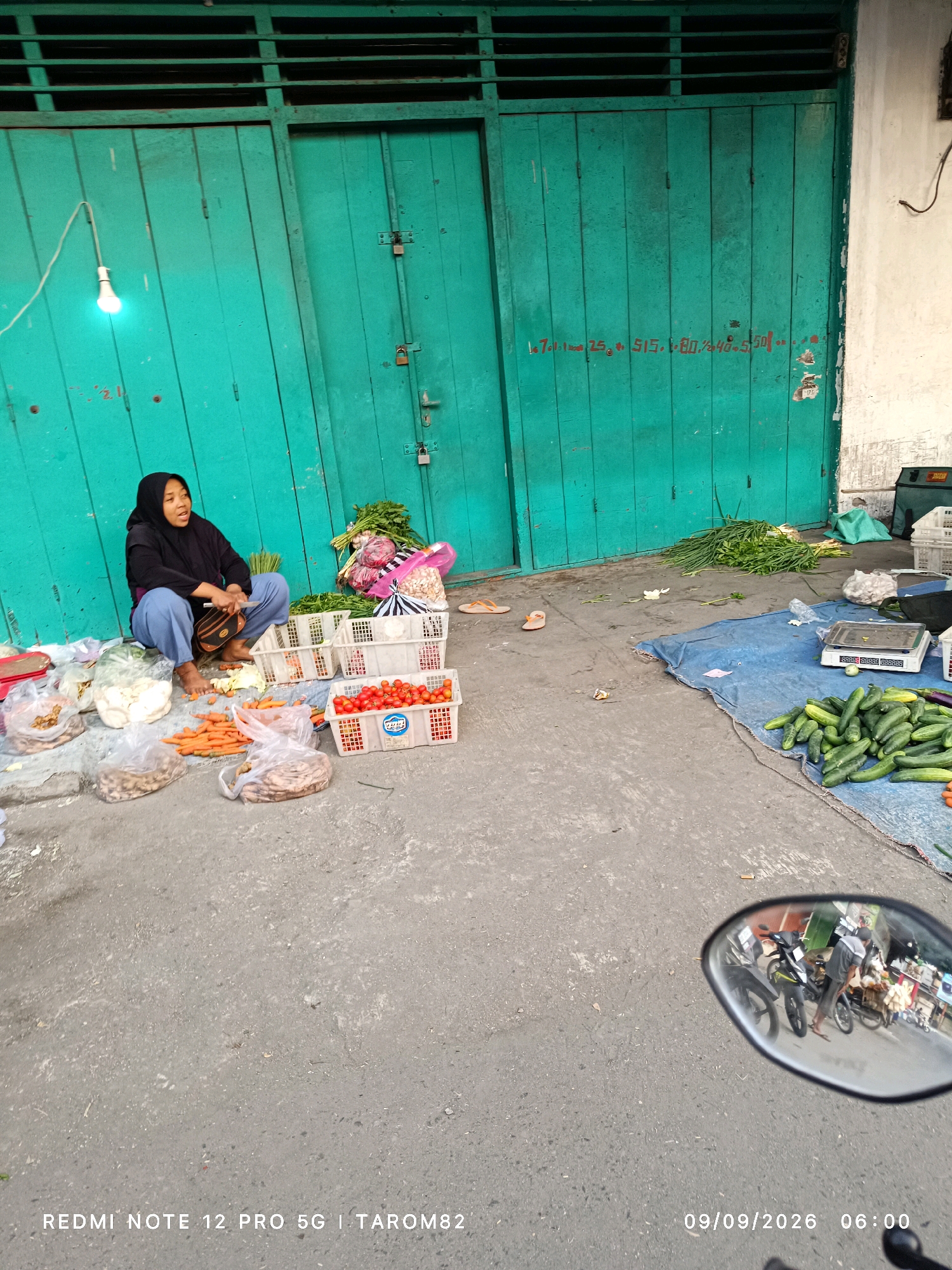 This image depicts a street-side market scene in Ngoro, Indonesia, at 6:00 AM on September 9, 2026. A woman wearing a hijab and blue pants sits on the ground, surrounded by various fresh produce. Baskets of tomatoes and other vegetables are visible, along with plastic bags containing ginger or similar root vegetables and loose carrots. In the background, a large, teal-colored door with several padlocks serves as a backdrop, and a bare light bulb hangs from the top left. To the right, a blue tarp is spread on the ground, displaying a digital scale, a pile of cucumbers, and more vegetables. The reflection in a motorcycle's rearview mirror shows a man attending to a cart of goods and other motorcycles. The overall environment is an outdoor urban setting with a concrete ground.
