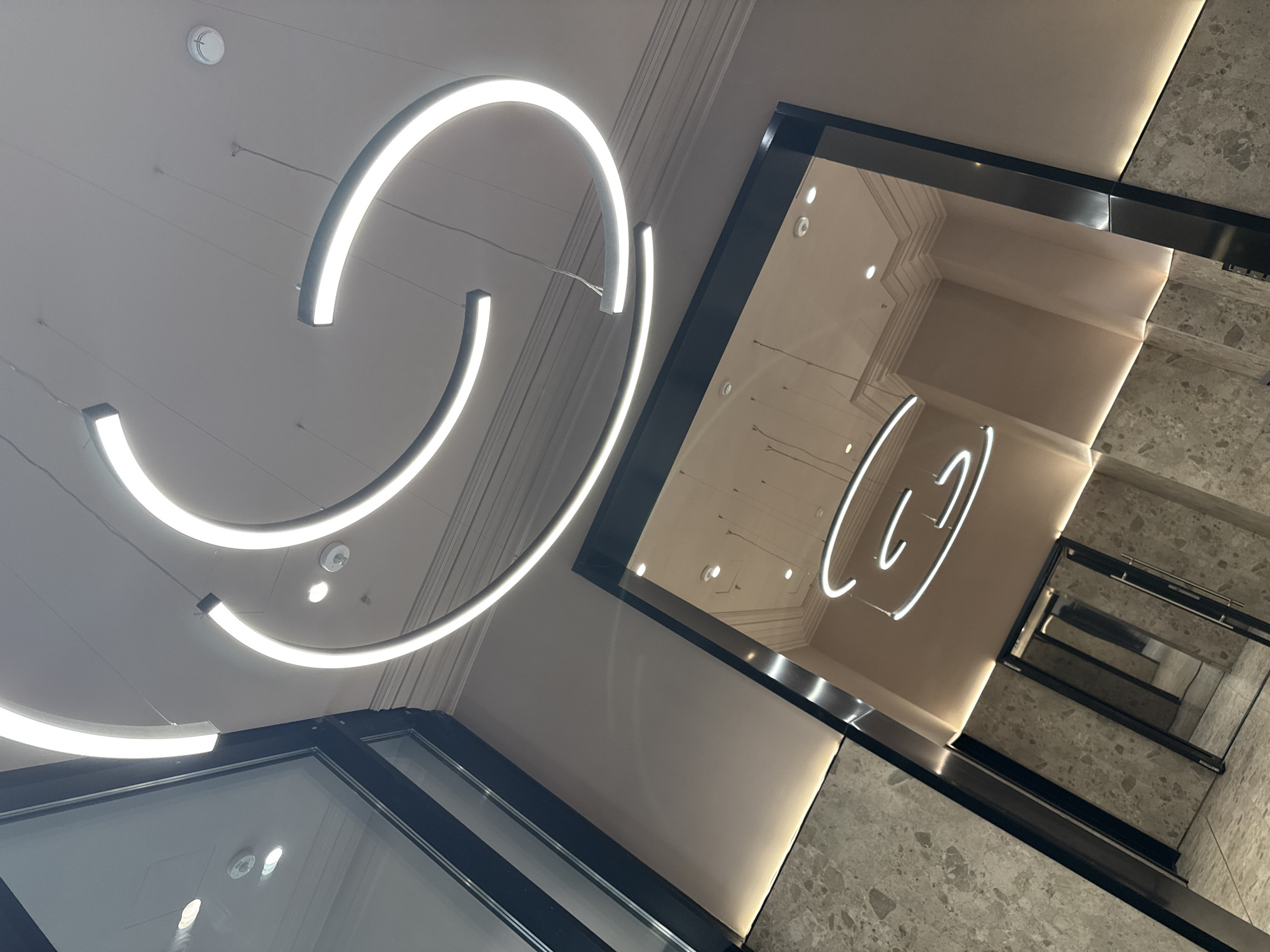 A wide-angle, low-angle shot captures the interior of a modern building. The scene is dominated by a ceiling with three large, curved, white LED lights, arranged in concentric arcs. The ceiling is painted a light neutral color and features recessed lighting fixtures and what appear to be smoke detectors.
The lower portion of the image reveals a reflected scene in a mirrored surface, likely a large glass panel or door. This reflection shows a hallway or lobby area with marble-patterned walls and floor. The reflection also shows architectural details like crown molding and what seem to be doorways or openings.
The overall aesthetic suggests a contemporary, clean, and upscale environment, possibly a hotel lobby, office building, or high-end retail space. The lighting is bright and uniform, highlighting the sleek lines of the fixtures and architectural elements. The image composition draws the viewer's eye upward towards the dramatic lighting installation.