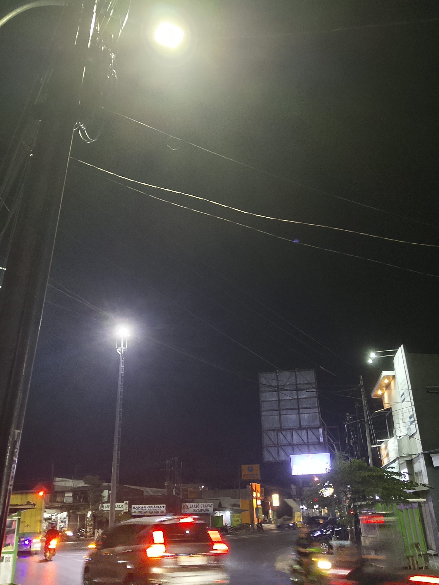 This is a night-time photograph taken from a street level perspective in Balun, Indonesia. The scene is dominated by a bright street light in the upper left quadrant of the image, casting a strong glow that illuminates a network of power lines stretching across the dark sky. Below the street light, a tall, slender lamppost with an illuminated head is visible.
The street itself is paved and appears to be a busy thoroughfare, with several vehicles in motion. A car with its brake lights illuminated is in the foreground, moving from left to right. To the left of the car, a motorcycle is also visible. In the background, there are various buildings, including what appear to be shops or small businesses with illuminated signs. One sign reads "BAKSO GRANAT KETAPANG." A large, unfinished billboard structure is also present, with a lit screen on it. Trees are visible alongside the road. The overall impression is of a typical urban street scene at night, illuminated by artificial lighting and busy with traffic.