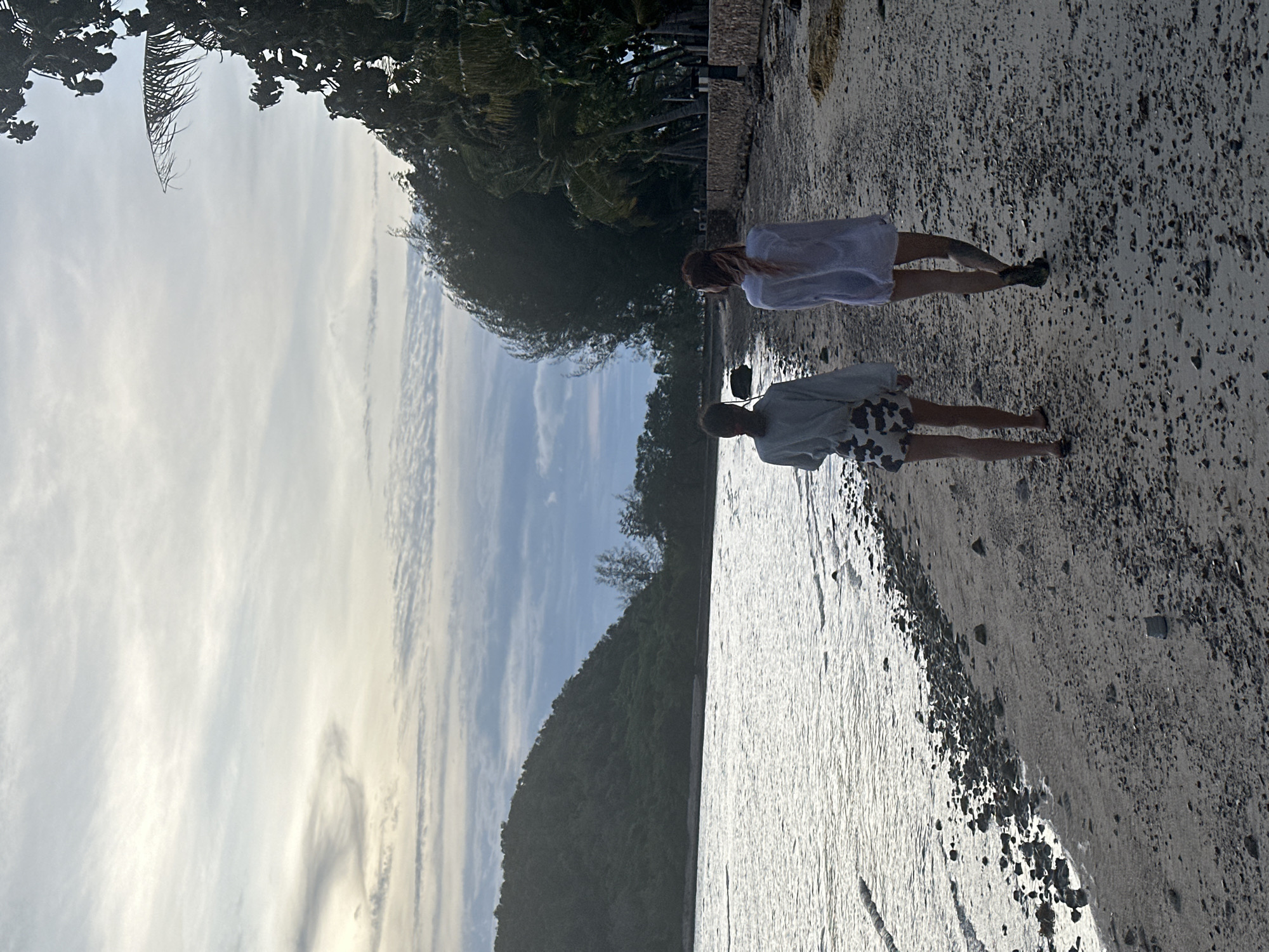 This is a vertical, full-shot image of two people walking along a sandy beach with a rocky shore. The person in the foreground is wearing a light-colored shirt and patterned shorts, while the person behind them is wearing a white dress. Both individuals appear to be walking away from the camera, towards a body of water that reflects the sky.
The scene is set on a beach in Ko Samui District, Thailand, indicated by the tropical foliage and palm trees visible in the background. The sky is partly cloudy, with wispy cirrus clouds, suggesting it might be late afternoon or early morning, with soft, diffused light. The water is calm, with gentle ripples on its surface. The sandy beach is dotted with small pebbles and rocks, and the waterline is clearly visible.
To the left of the frame, a densely vegetated hillside rises, covered in trees and lush greenery, indicating a tropical environment. The overall mood is serene and peaceful, suggesting a leisurely stroll along the coast. There is no visible text in the image.