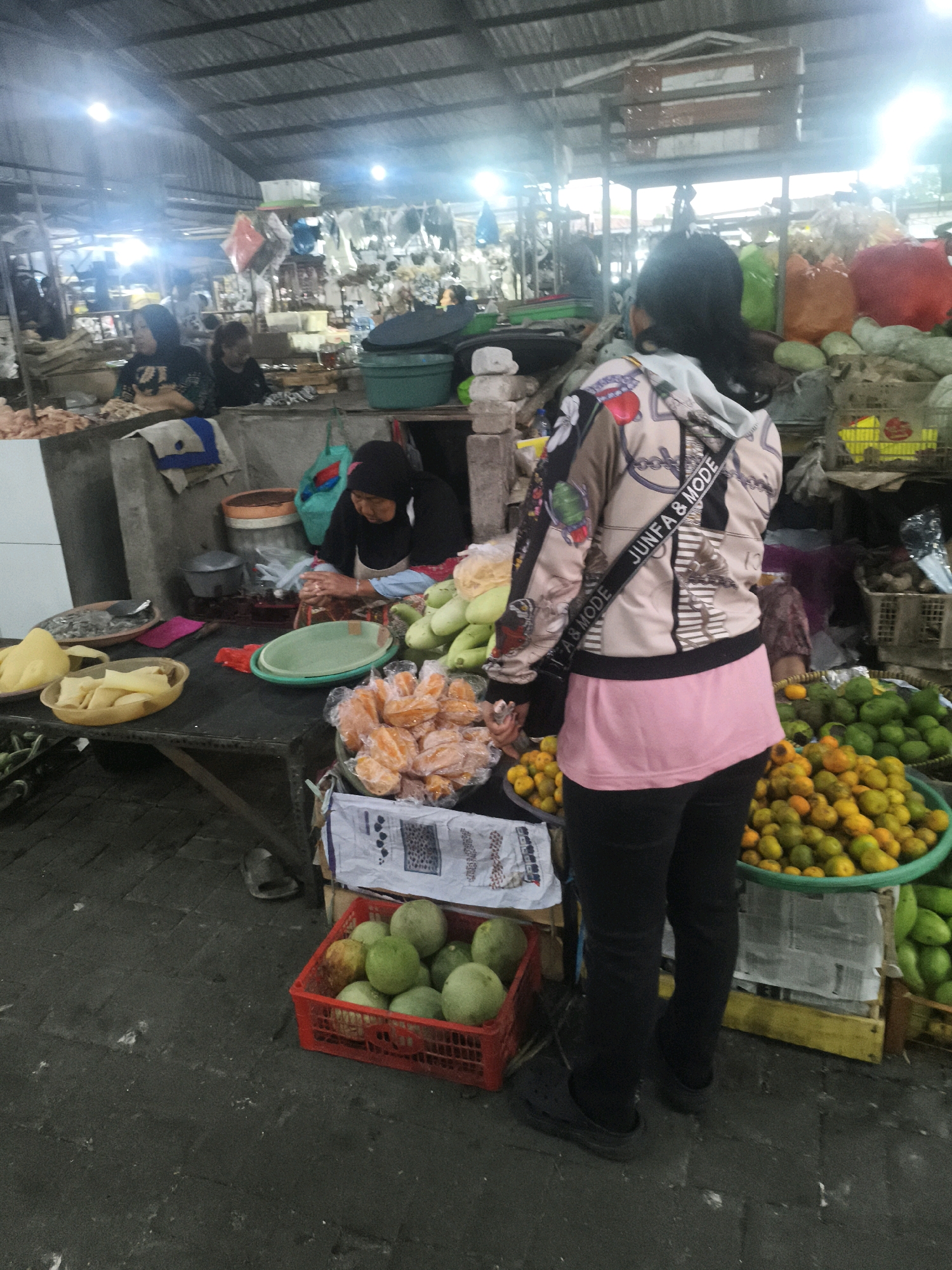 The image depicts a bustling indoor market scene, likely in Surabaya, Indonesia, characterized by its open-air structure with a corrugated metal roof and fluorescent lighting. In the foreground, a person stands with their back to the camera, wearing a patterned jacket over a pink top and black pants. They are carrying a bag with the text "JUNFA & MODE" on the strap.
To the left, a vendor wearing a black hijab and apron is seated behind a table laden with food items. She is actively engaged in preparing or displaying her goods, which include what appear to be slices of fruit or cheese in yellow bowls and a large quantity of orange-colored food items in a plastic bag. Other food items, such as green gourds and citrus fruits in a green basin, are visible on separate tables. Various baskets and containers are scattered throughout the scene, some holding produce and others appearing empty. The ground is paved with dark, textured material. The overall impression is one of lively commerce within a traditional market setting.