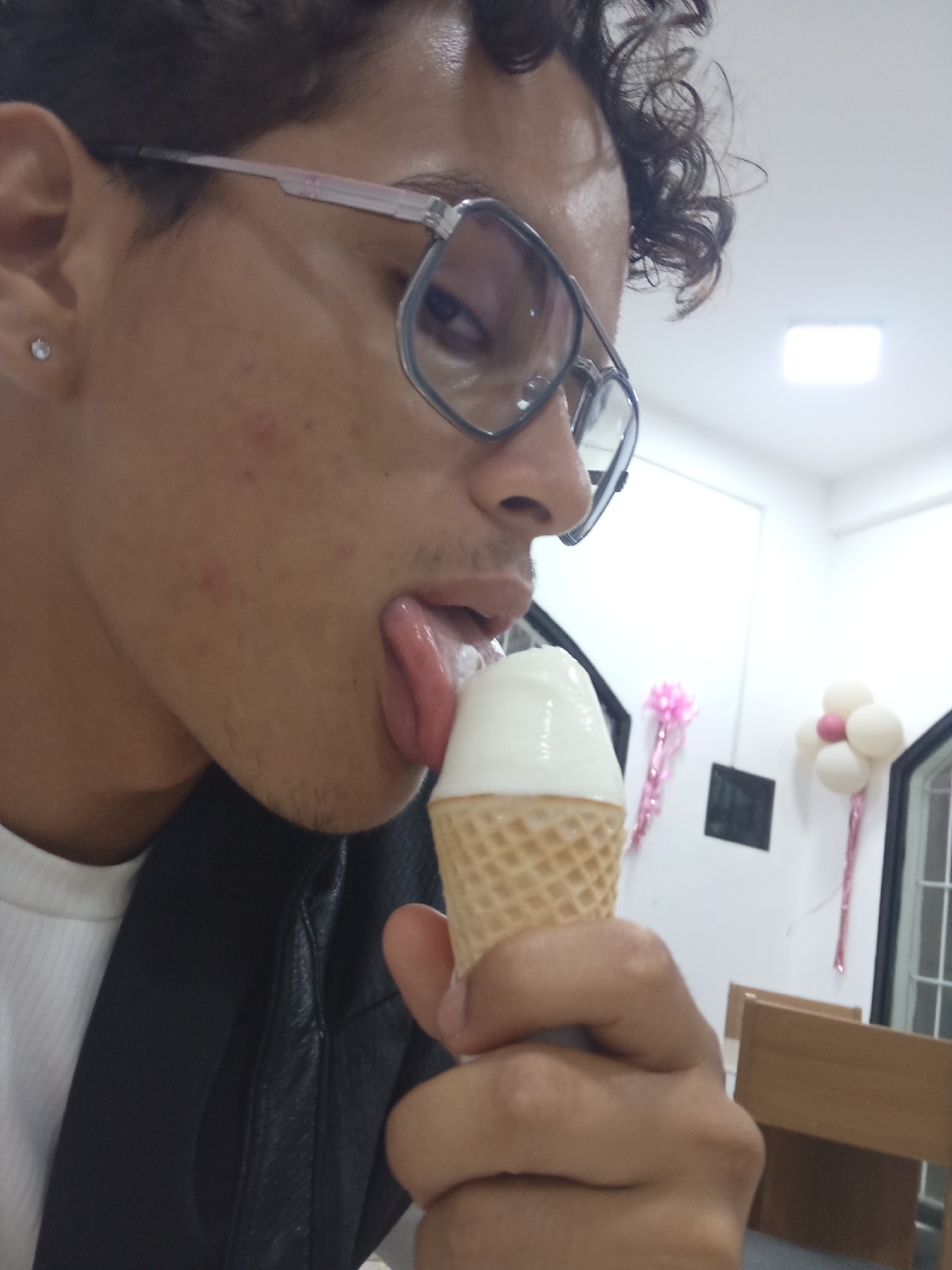 A person with dark curly hair and glasses is licking a vanilla ice cream cone. They are wearing a white shirt under a black jacket. The background appears to be an indoor space with white walls and decorations, possibly for a party or celebration, including a balloon arrangement resembling a flower. A brown wooden chair is partially visible. The location is not discernible from the image. The city of Juan Griego, Venezuela cannot be confirmed.