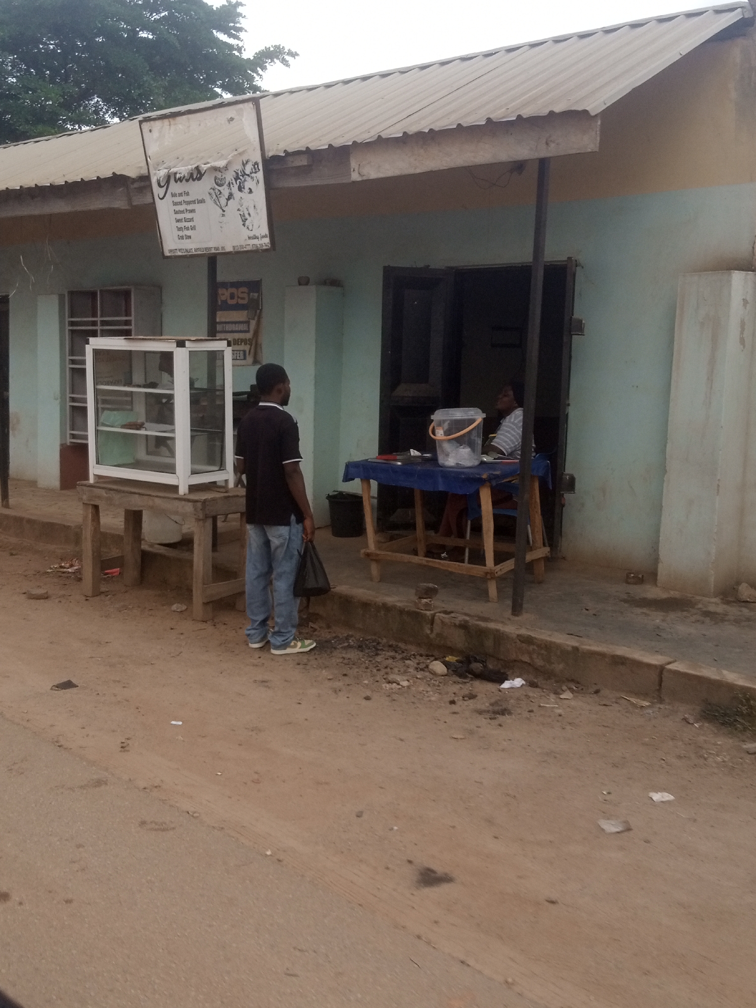 In Gura Top, Nigeria, a modest storefront features a corrugated metal roof extending over a light blue, two-story building. To the left, a white display case sits atop a wooden table. Two men stand in front of the building, one facing away from the camera. A third person is seated inside the establishment behind a table. The ground in front of the building is dirt and gravel, with some scattered debris. A sign hangs from the roof, partially obscured.