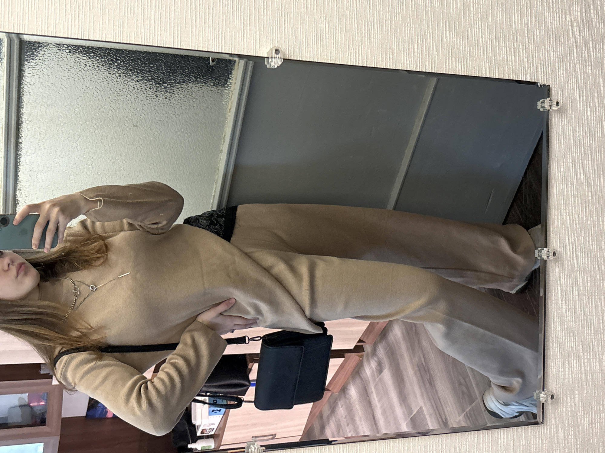A female figure in a beige sweater and pants is captured in a mirror selfie. She holds a smartphone in her left hand, partially obscuring her face. A black purse hangs on her left side. The mirror is framed with hardware, reflecting a room with cabinets and a textured wall. The location appears to be indoors, but there's no clear indication that the setting is in Saint Petersburg, Russia.