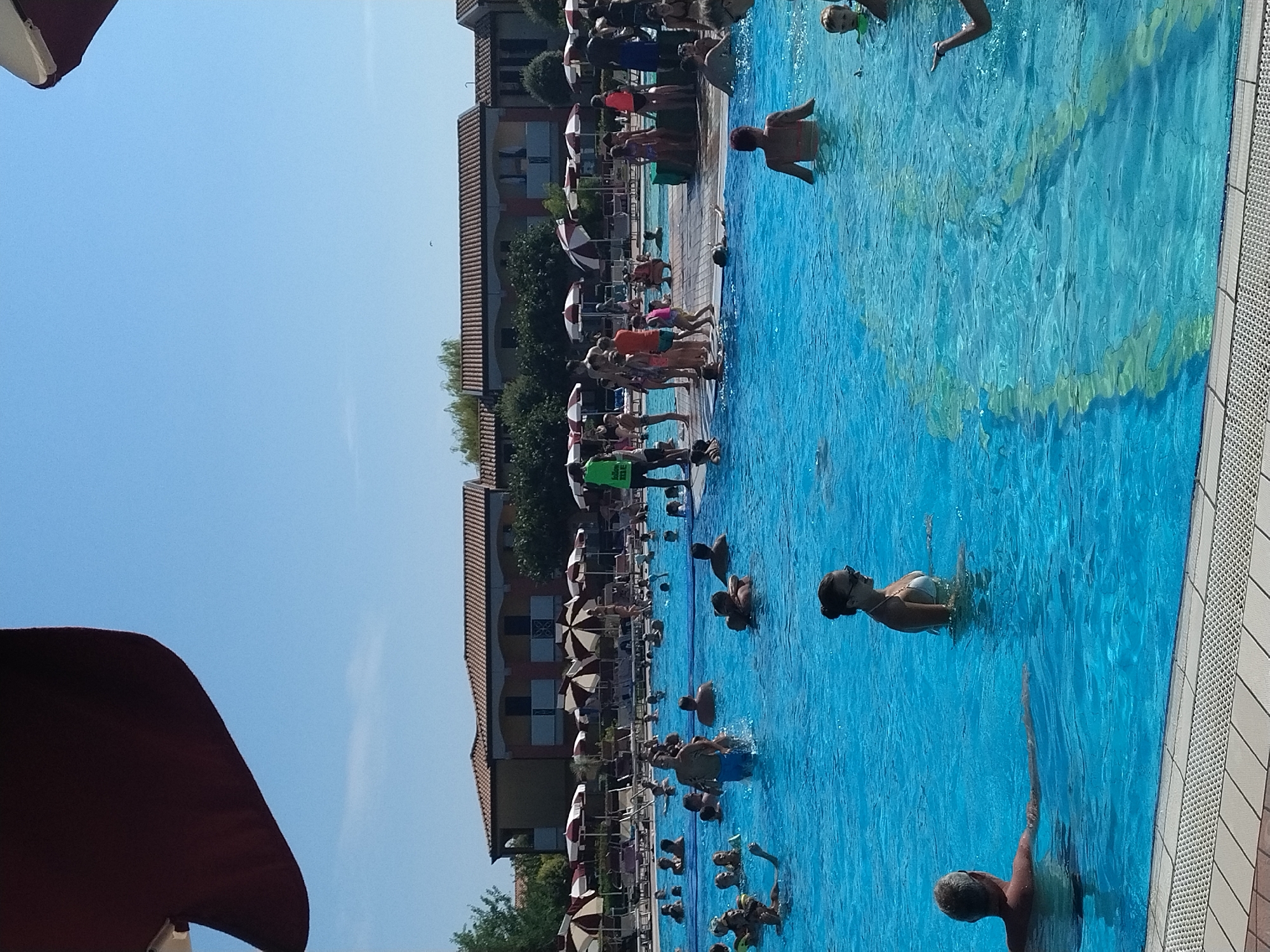 The image shows a crowded outdoor swimming pool on a sunny day. The pool is filled with blue water and many people of various ages are swimming, wading, and standing in the water. Some individuals are partially submerged while others are fully immersed. In the foreground, a man with grey hair is visible in the water, his arm extended. To his right, a woman with dark hair in a white bikini is also in the water, looking to her right. Further in the background, a lifeguard in a green vest with "FOLLOW ME" written on it is standing near the edge of the pool. Umbrellas and lounge chairs are set up around the pool area. Buildings with terracotta roofs are visible behind the pool. The scene appears to be a leisure resort or water park. The presence of umbrellas, lounge chairs, and the overall atmosphere suggests a resort in Caorle, Italy, known for its family-friendly attractions and swimming pools.