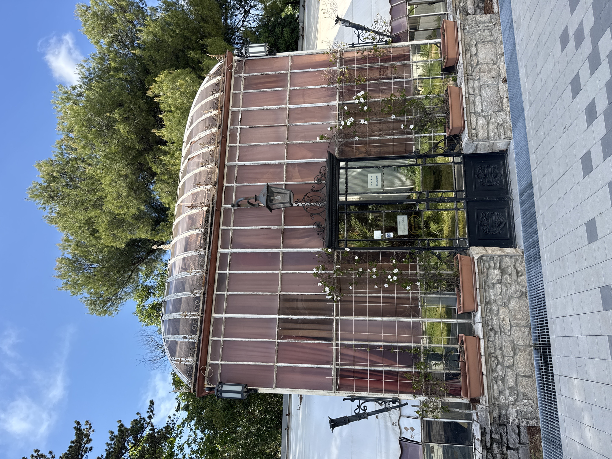 A sideways shot reveals a historical structure identified as the "Kraljičina bašta (Queen's garden)," located in the city of Bar, Montenegro. The structure is characterized by a glass-enclosed facade and roof. The building's entrance is framed by a black wrought-iron gate, adorned with decorative patterns. White flowering vines climb the facade. The building's base is constructed with rough-hewn stone. The structure abuts a paved area and is partially obscured by mature trees. Street lamps are affixed to the building. The sky is a clear blue with light clouds.
