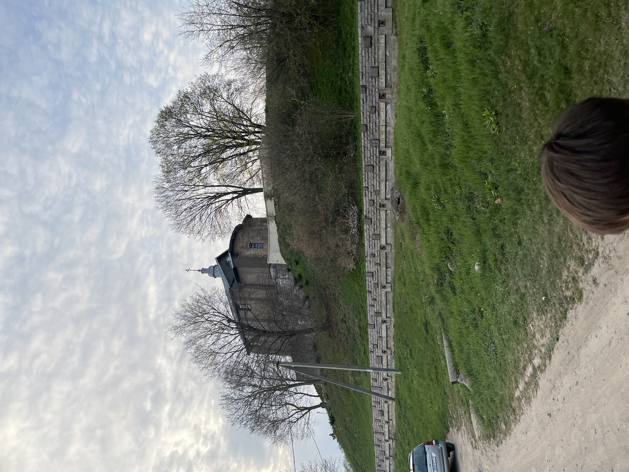 The image depicts a stone church building situated on a raised area, partially obscured by bare trees. A gray sky filled with scattered clouds forms the backdrop. Adjacent to the church's hill is a grass-covered area bordered by a structured stone wall. A sandy path runs along the bottom right of the image, with a silver car visible parked near the edge of the path. Part of a person's head is visible in the upper right corner. The location is Unknown, Unknown.