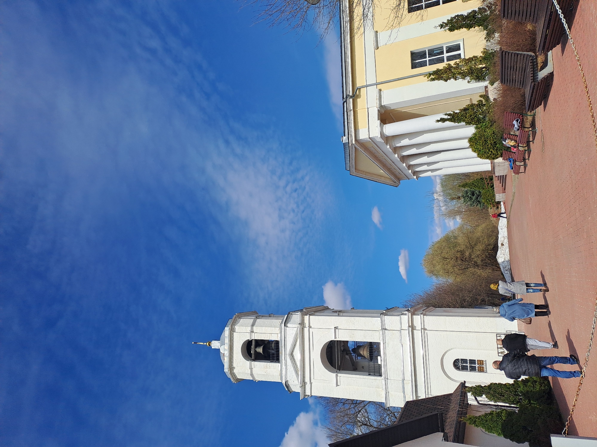 A low-angle shot, rotated counterclockwise, features two buildings under a blue sky with cirrus clouds. A white bell tower dominates the foreground, complete with visible bells and arched openings. Several individuals are present; some are walking near the tower, while others are seated on benches adjacent to a yellow building with white columns. The ground appears to be paved with red bricks, and landscaping includes trees and shrubs. A chain serves as a border. This is the Ivashevo, Russia.