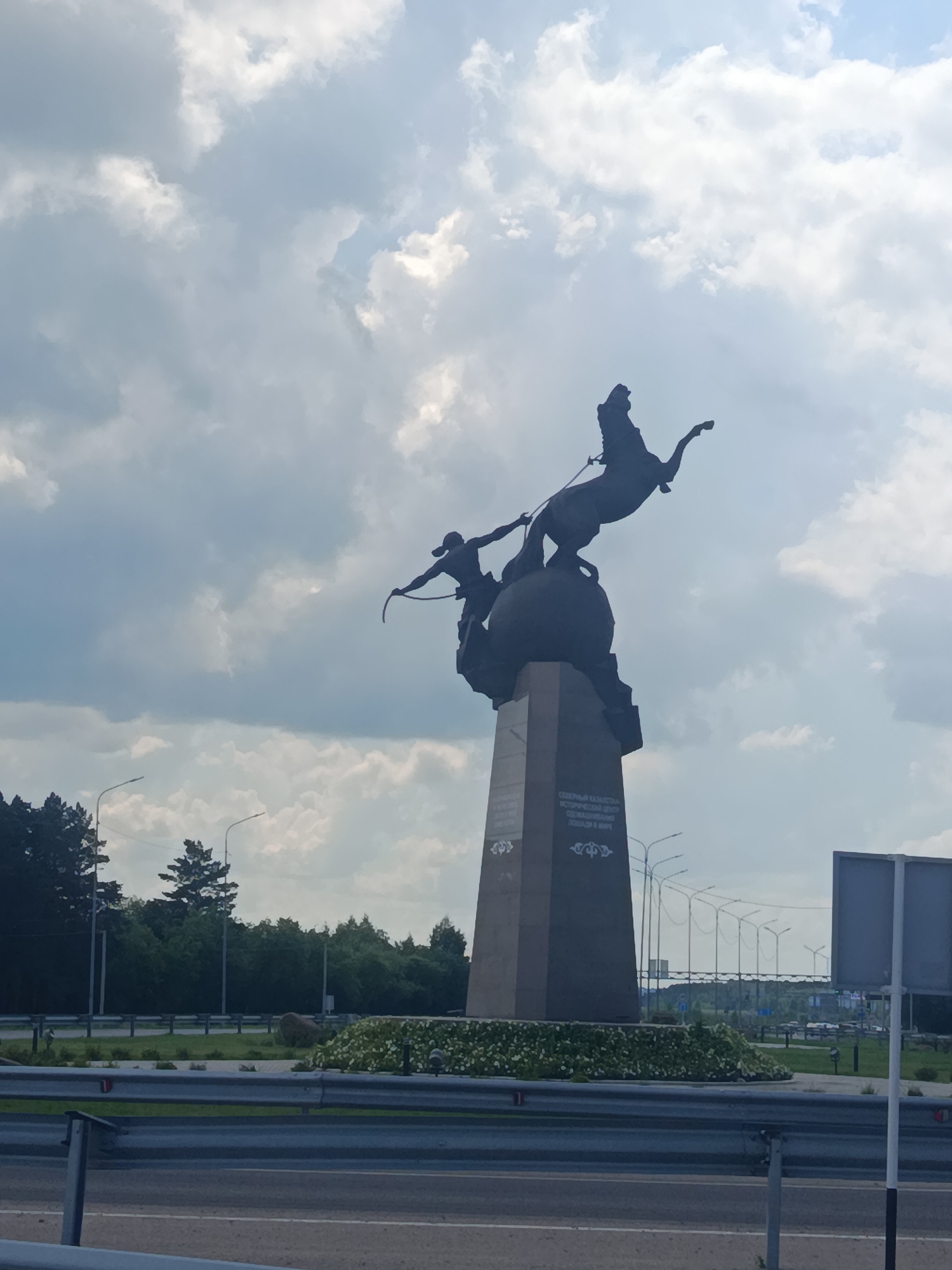 A monumental sculpture of a horse and rider is silhouetted against a cloudy sky. The sculpture, made of dark material, depicts a horse rearing up on its hind legs, with a rider holding a bow and arrow in pursuit. The base of the sculpture is a large, tapered obelisk, also dark in color, with some inscription visible on its surface. Around the base, a low, circular garden bed is filled with white flowers. The sculpture is situated in an outdoor urban environment, likely a roundabout or park, with a road and metal guardrails in the foreground. Trees and streetlights are visible in the background, suggesting a public space. The sky is filled with large, dramatic clouds, hinting at either an overcast day or the approach of a storm.