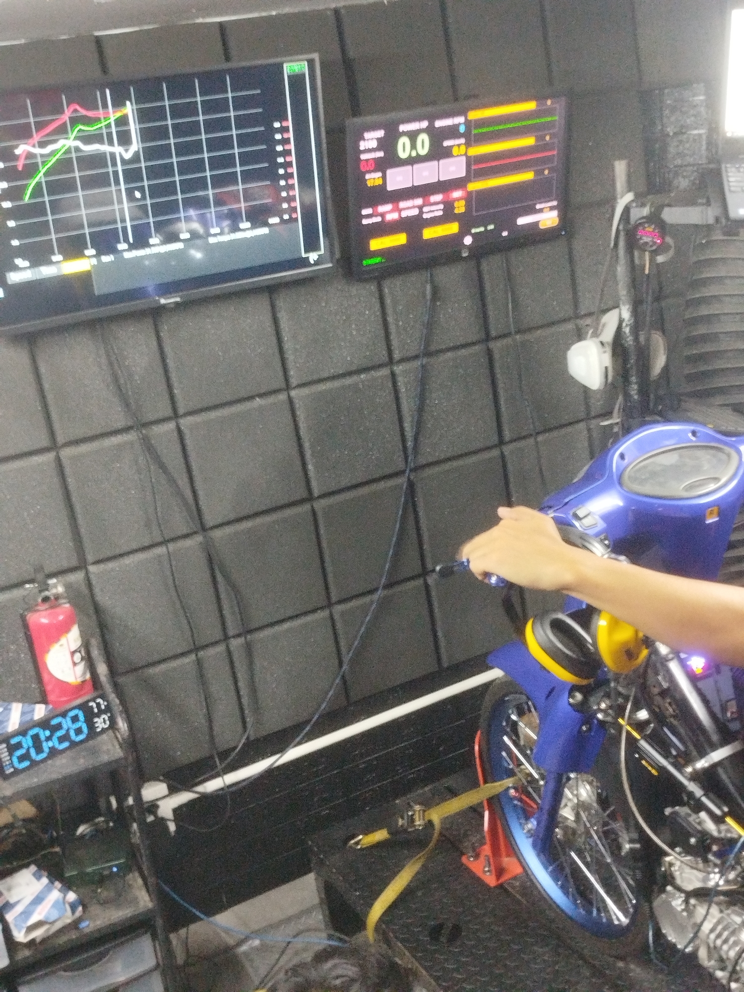The image depicts a dynamometer testing setup for a motorcycle, likely in a workshop environment in Ban Kho En, Thailand. A blue scooter is positioned on the dynamometer, its rear wheel connected to the testing apparatus. A person's hand is visible on the handlebars, suggesting active operation. Two screens are prominently displayed: one on the left shows performance graphs with multiple colored lines, while the screen on the right displays digital readings and bar graphs, with "0.0" being a prominent figure. To the left of the screens, a red fire extinguisher is mounted on a sound-dampened wall, composed of square, textured panels. Below the screens, a digital clock displays "20:28" and "30," along with other illegible numbers. The overall impression is of a technical setup for measuring and analyzing motorcycle performance.