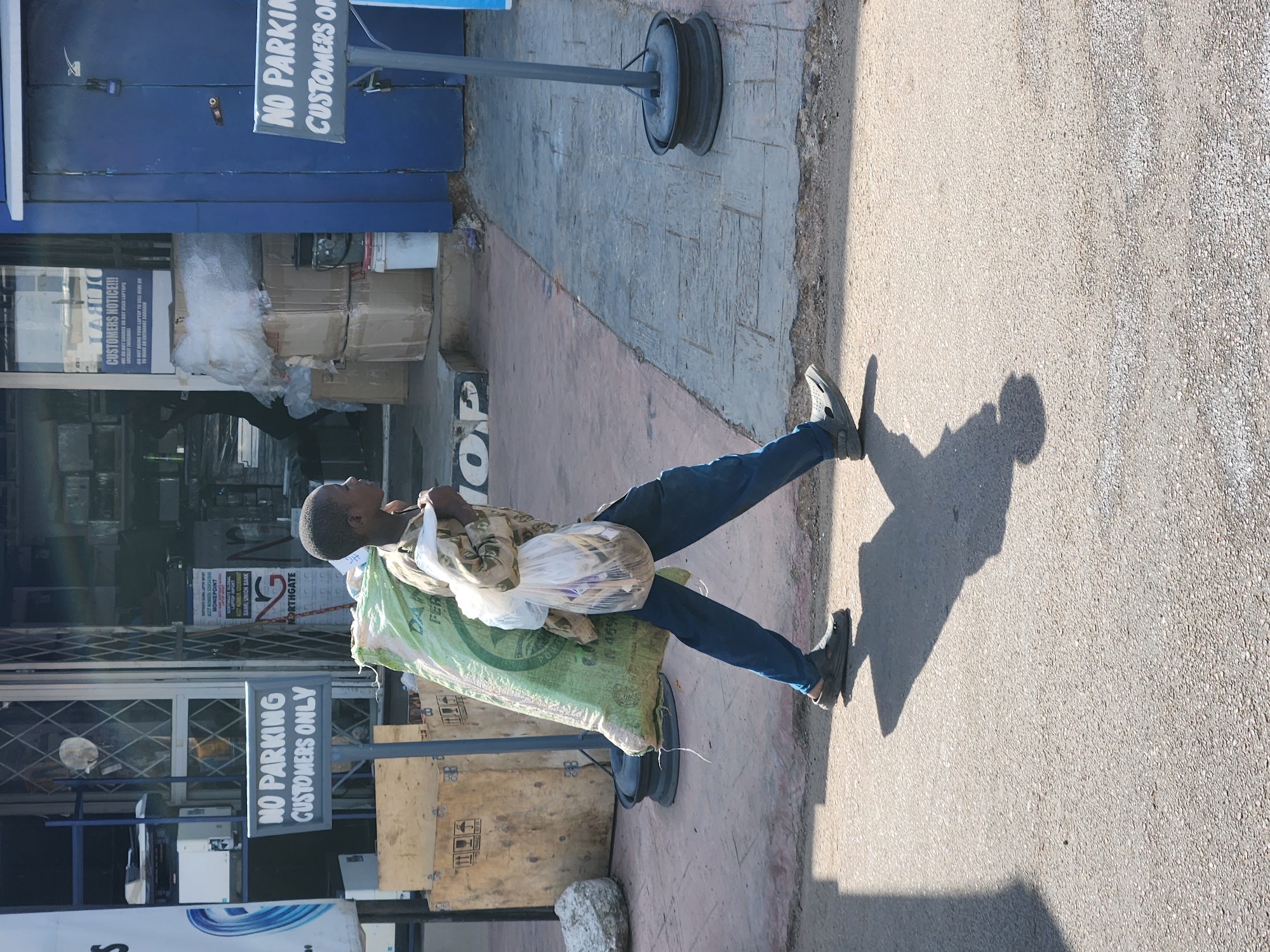 An individual is seen carrying items in the city of Unknown, Unknown. The person is holding a plastic bag and a green sack, standing next to a "No Parking Customers Only" sign. The background shows store fronts with signs and parked items. The scene is captured in daylight.
