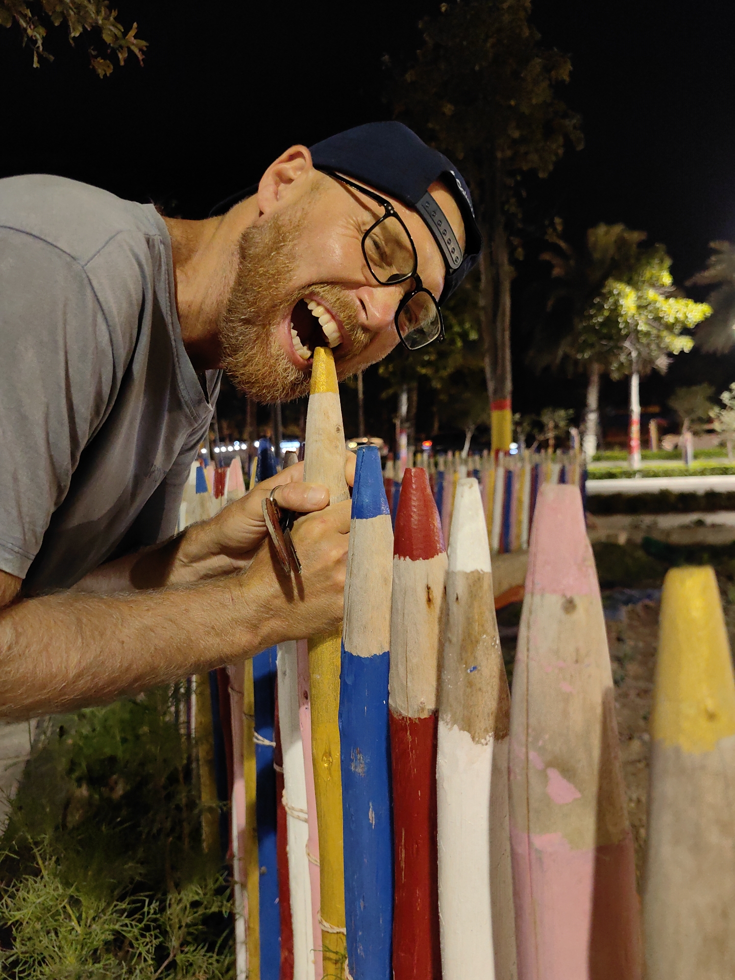 A man with a beard and glasses is biting the yellow top of a fence made of large, painted wooden pencils. The fence is made of colored pencils and is in Bai Chay Ward, Vietnam. He is wearing a gray t-shirt and a dark baseball cap. He is holding a set of keys in his left hand. The background is dark, with trees and streetlights visible.
