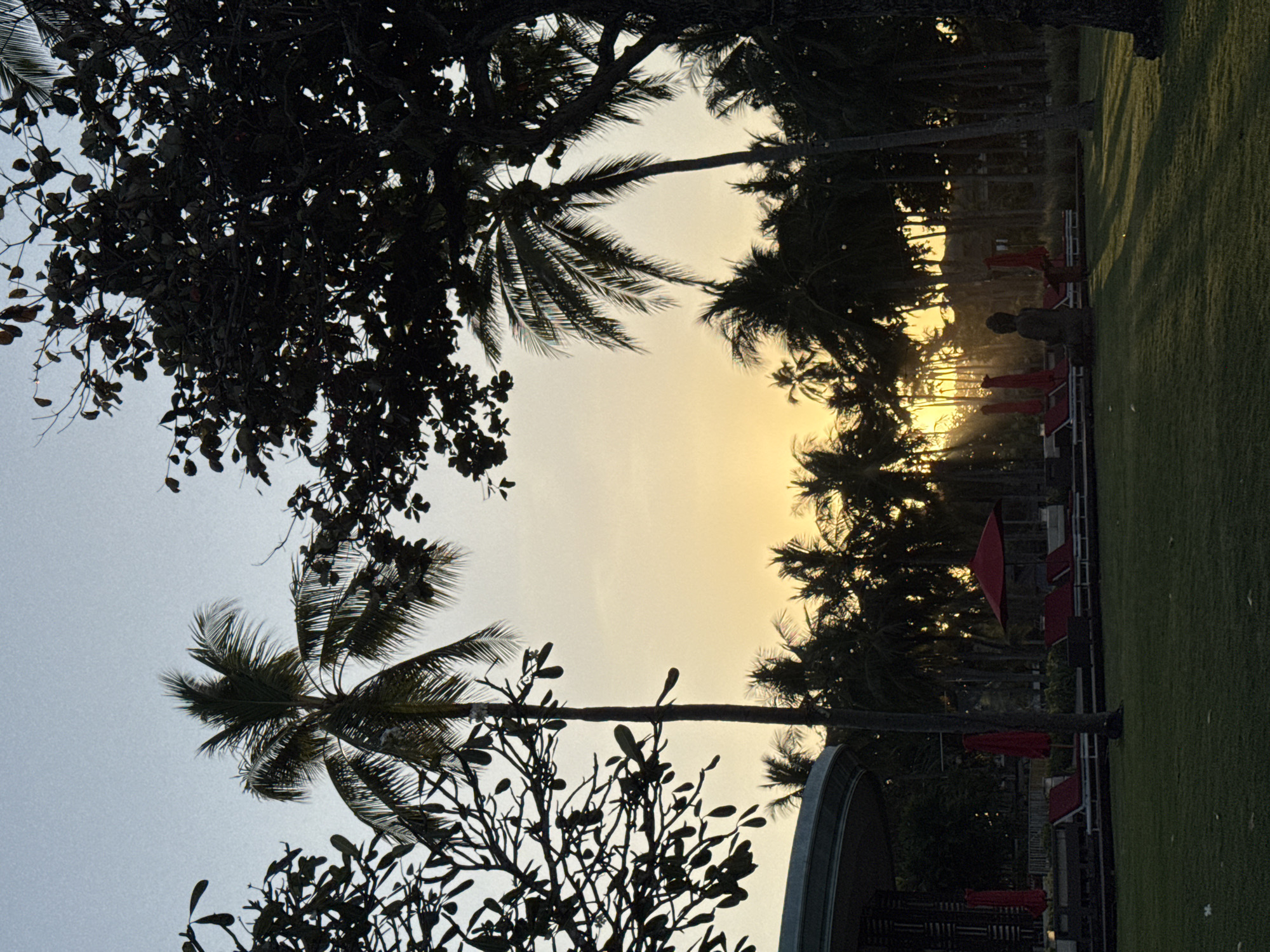 A serene, silhouetted scene unfolds in Kuta Selatan, Indonesia, at sunset. The sky is a gradient of soft blues and yellows, with rays of sunlight piercing through the fronds of numerous palm trees. In the foreground, a well-maintained green lawn is cast in long shadows from the trees. A dark, seated Buddha statue is positioned on the lawn. Further back, a row of red lounge chairs and umbrellas are arranged around a pool area, with a modern building structure to the left. The image captures a peaceful, resort-like atmosphere during the golden hour.