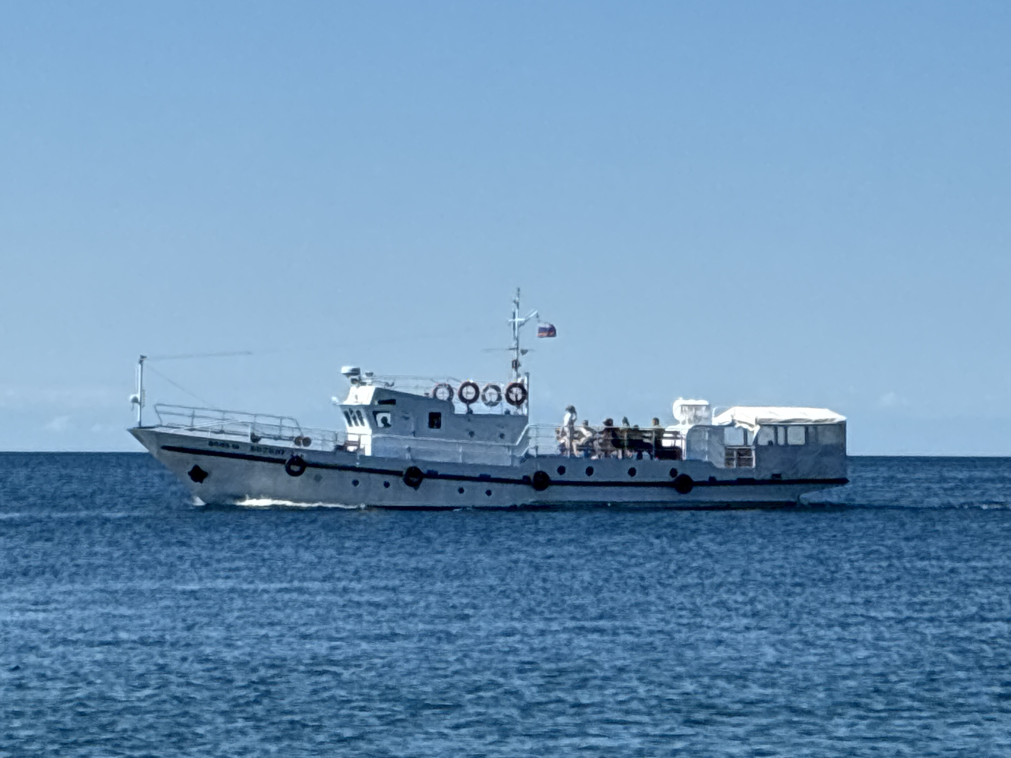 A white tourist boat is cruising on the blue waters of Lake Baikal under a clear sky. The boat has a cabin structure at the rear with windows and a white canvas roof. Several people are visible on the deck of the boat, some standing and others seated. The boat has a white railing along its sides and several lifebuoys are mounted on the upper deck. The name "BOSS 10" is visible on the hull of the boat. The scene is likely located in the vicinity of Listvyanka, Russia, a popular tourist destination on the western shore of Lake Baikal, known for its boat tours. The water is slightly choppy, indicating the boat is in motion.