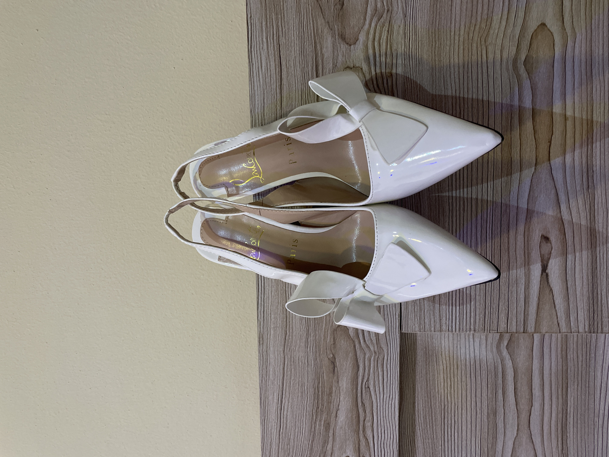 The image shows two white patent leather slingback heels with bows resting on a surface with a light-colored wood grain pattern. Part of a light yellow wall is visible in the background to the left of the wood surface. There is no indication of location in the city of Bar Jirgi Summa, Nigeria.