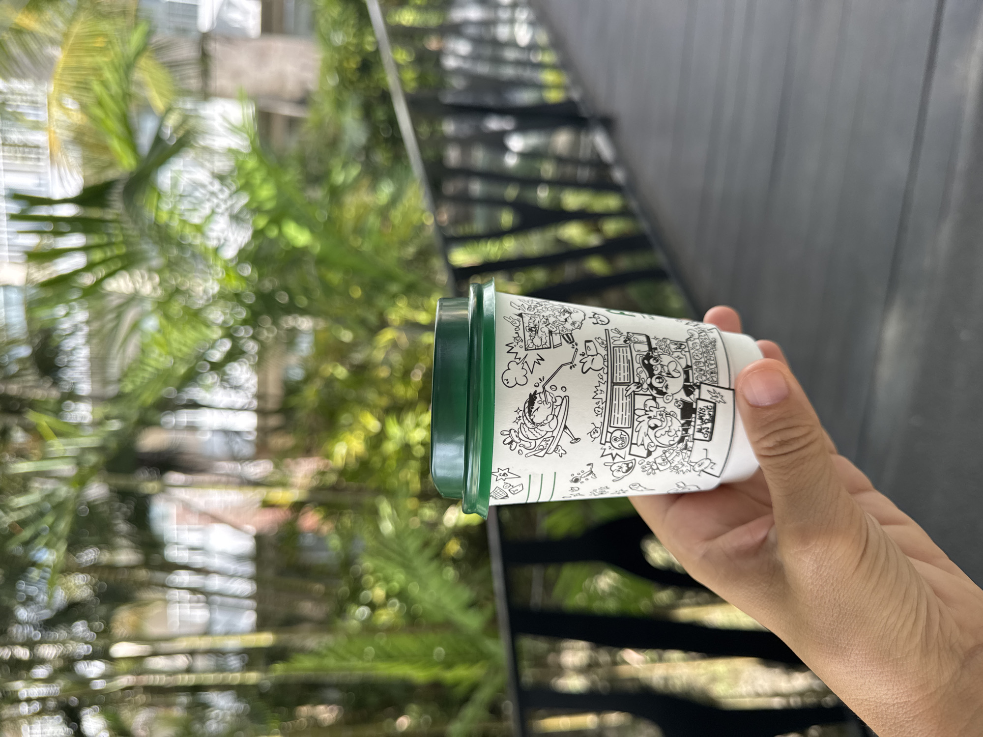 A person is holding a white paper cup with black cartoon drawings and a green lid in their right hand. The background includes a black metal structure, likely a staircase or railing, as well as blurred green foliage, suggesting an outdoor or garden setting. Given the prompt's request to provide the location of Kuta Selatan, Indonesia, it's presumed that this photograph was taken outdoors, perhaps in a garden or near a building with greenery in Kuta Selatan. The absence of specifics like a visible building or a particular type of plant restricts further detailed description of the environment. There are no people visible other than the hand holding the cup. The action is limited to holding the cup; any interaction is absent.