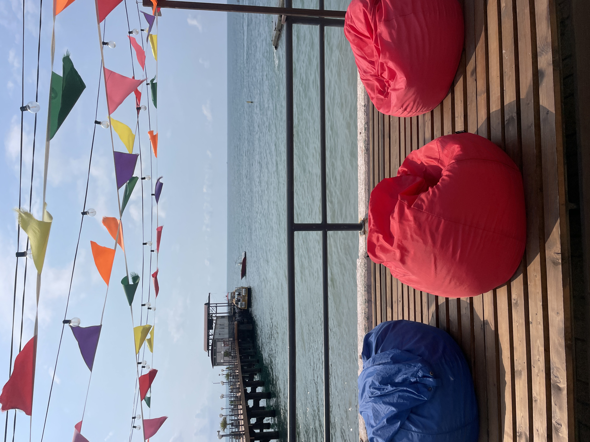 This is a photograph of a waterfront seating area, possibly in Sochi, Russia. In the foreground, three large, colorful beanbag chairs are arranged on a wooden deck. The chairs are blue, red, and another red one. A black metal railing separates the deck from the sea beyond. In the mid-ground, a wooden pier extends into the water. At the end of the pier, there is a building with a covered roof and several windows. A yellow boat is docked next to the pier, and a red boat is in the water nearby. The sea is calm and stretches out to the horizon under a partly cloudy sky. Colorful triangular flags and string lights are strung across the top of the image, adding a festive atmosphere. The overall impression is one of a relaxed outdoor seating area by the sea.