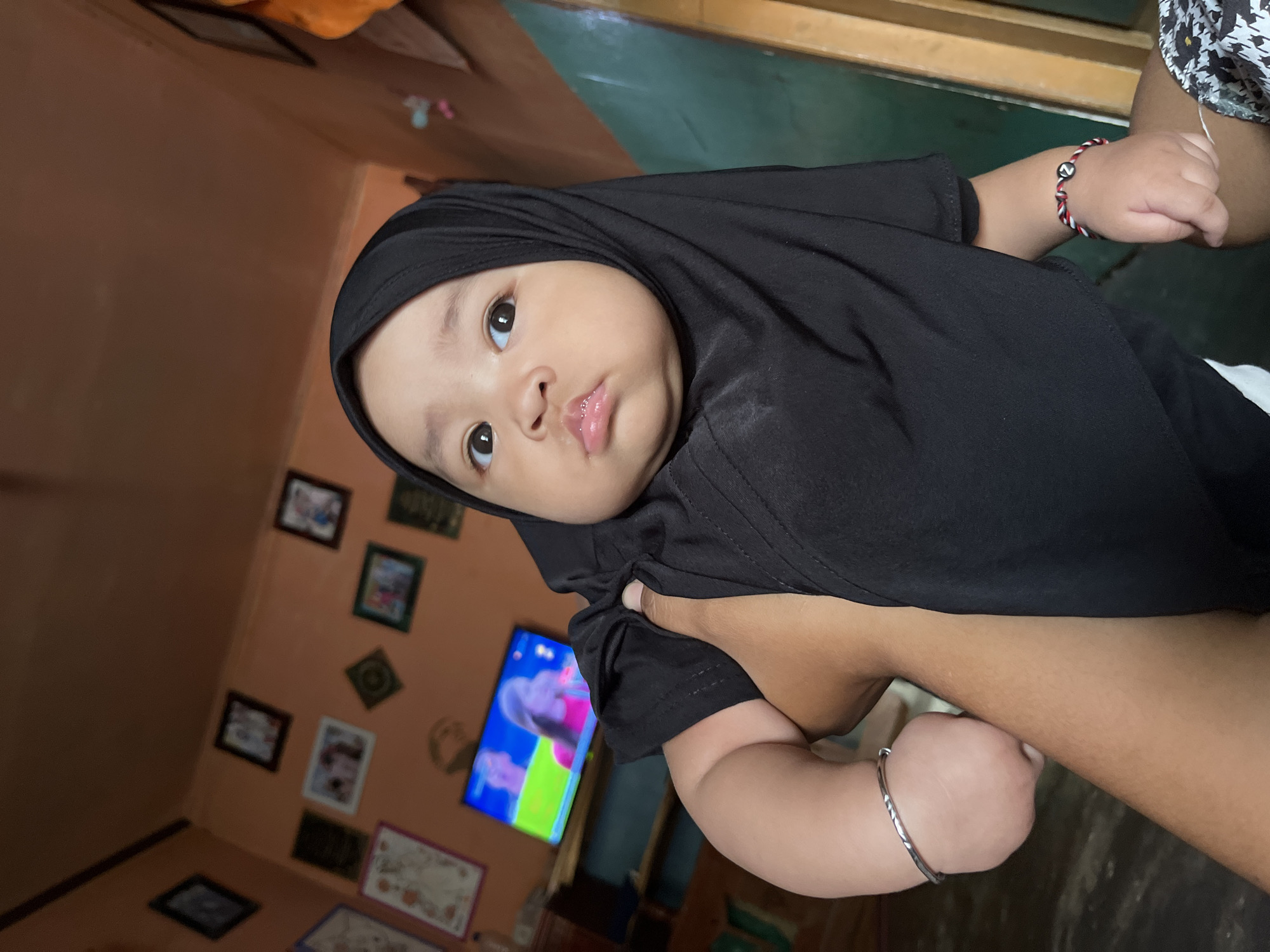 A baby wearing a black hijab is being held by an adult. The baby has big, dark eyes and full cheeks. The adult's arm is visible holding the baby. In the background, there is a television displaying a colorful image, framed pictures on the wall, and the room appears to be a home interior. The location is identified as Kruengmate, Indonesia.
