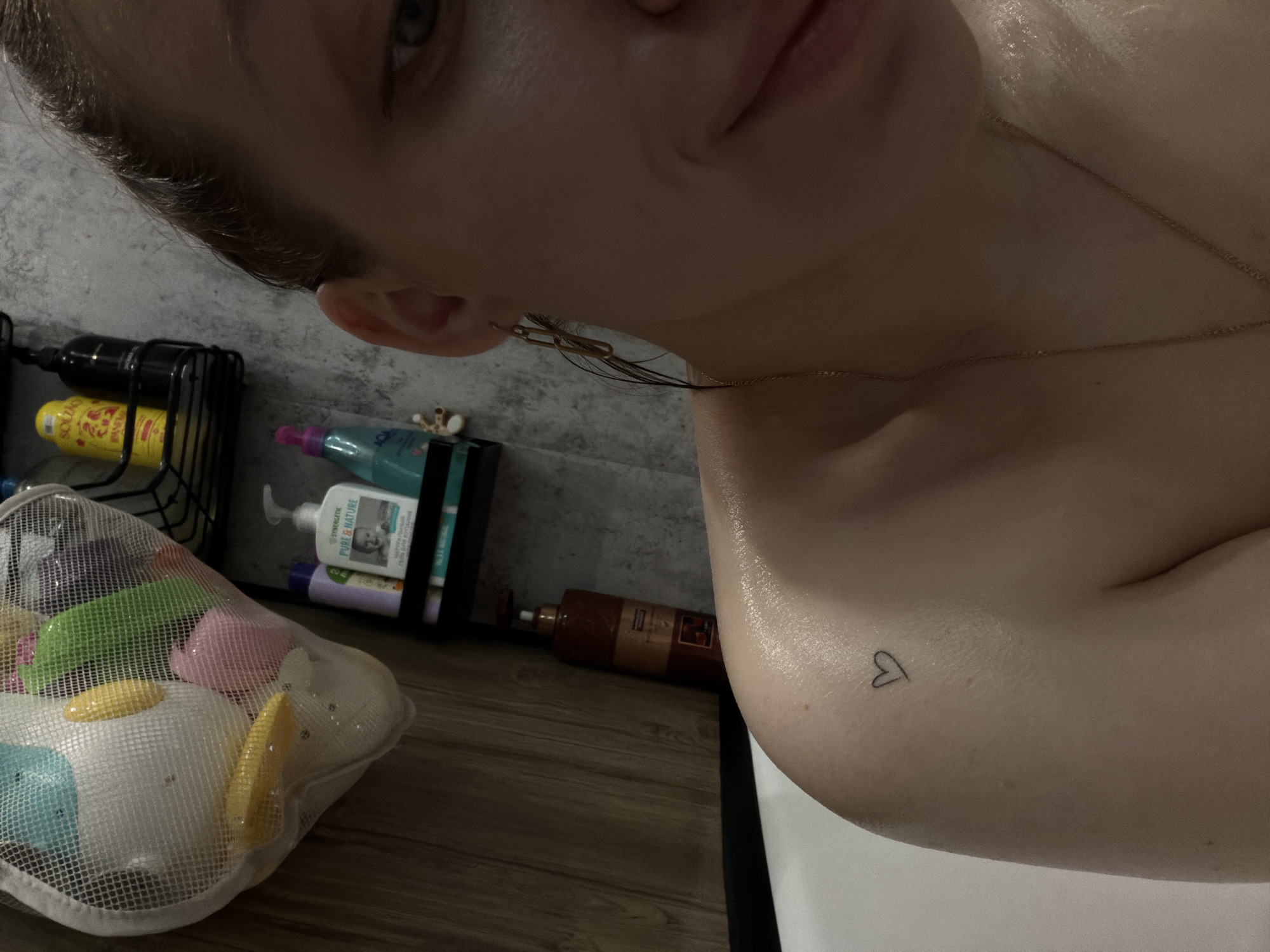 A close-up, high-angle photograph features a person from the shoulders up, focusing on their face and shoulder. The person has fair skin, wet hair, and is wearing a thin gold necklace. On their upper left shoulder is a small, minimalist heart tattoo. In the background, various bathroom items are visible. A black wire rack holds several bottles, including one yellow bottle and another that is black. A white mesh bag filled with colorful bath toys sits on the floor. Other bathroom products are also visible behind the mesh bag. The scene appears to be in a bathroom. There is no information to indicate that the location is in the city of Reshetnikovo, Russia.