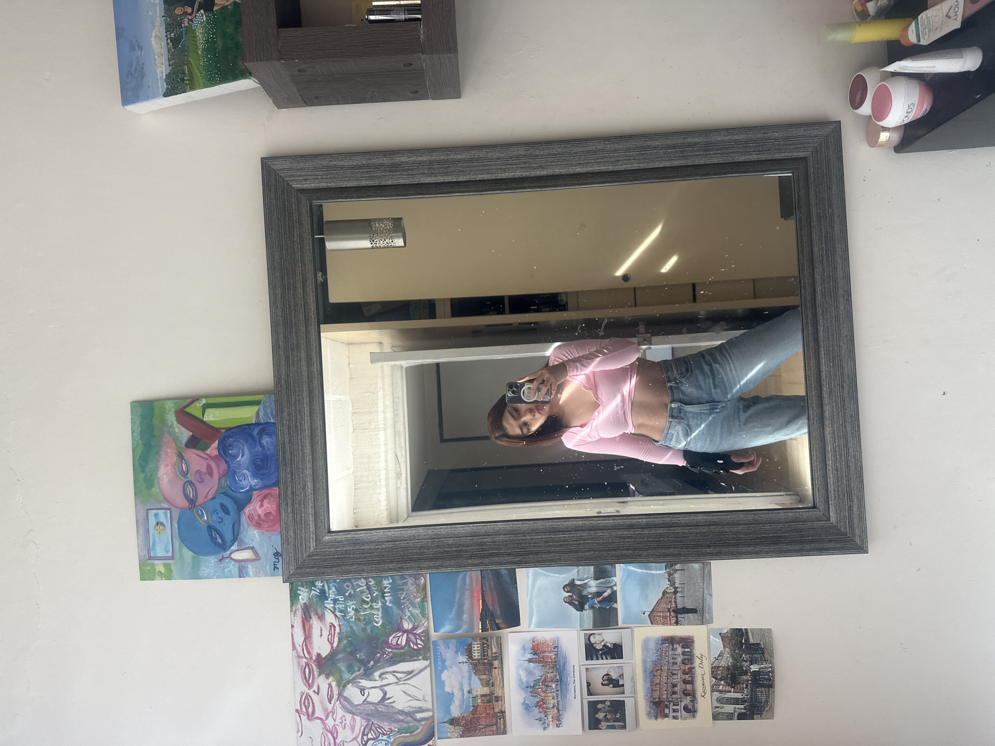 This media file captures a mirror selfie taken in what appears to be a personal room. The image, originally rotated, shows a rectangular mirror with a dark, textured frame mounted on a white wall. Reflected in the mirror is a young person with long, dark hair, wearing a pink long-sleeved crop top and light-wash blue jeans, holding a smartphone to capture their reflection. Their expression is neutral.
Around the mirror, the wall is adorned with various personal items. To the upper left (when viewed upright), a small shelf holds a painting depicting a green landscape with a blue sky. To the upper right, another shelf displays several cosmetic products, with visible brand names like "FONDS" and "VICHY." A colorful painting featuring stylized faces in blue, pink, and red, wearing sunglasses, is positioned to the left of the mirror.
Below the mirror, the wall is covered with a collage of postcards and small prints. These include urban landscape scenes, with text identifying locations such as "Warszawa" and "Kazimierz Dolny," indicating places in Poland. Interspersed are black-and-white photos, possibly polaroids, showing people. A larger, more abstract painting below the mirror features a face surrounded by floral elements, with the text "The things I did JUST SO I could make you MINE."
The scene is brightly lit, suggesting daytime, with light reflections visible within the mirror. While the user provides "Puebla, Mexico" as context, there are no specific visual cues within the image to explicitly confirm this location. The overall setting conveys a lived-in, personal space decorated with art and mementos.