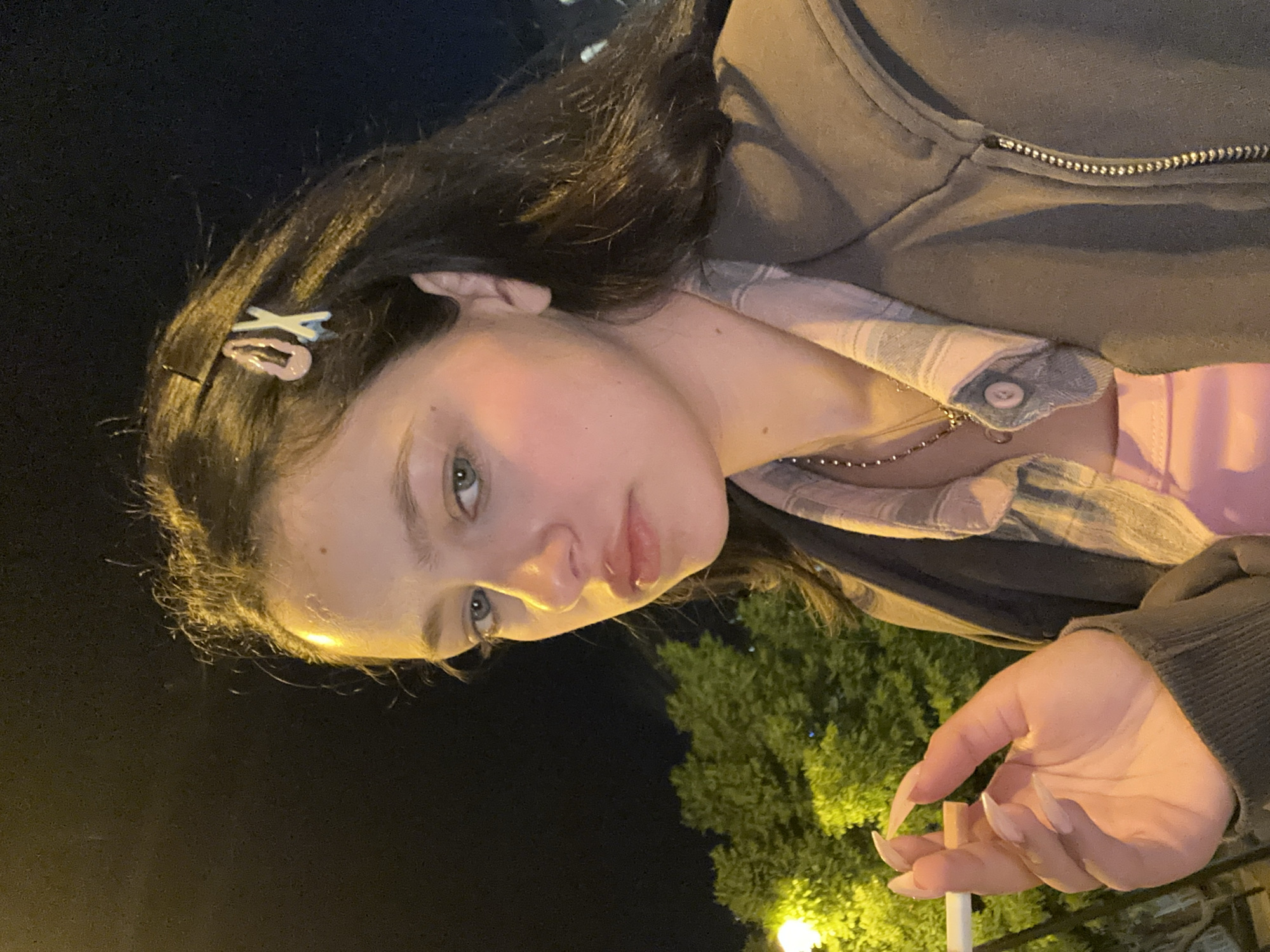 The image is a vertically oriented medium shot of a young woman outdoors at night, likely in an urban environment. She is fair-skinned with dark hair, accessorized with a hair clip, and is wearing a collared shirt and a zip-up jacket. She is holding a cigarette in her right hand. The background includes a tree illuminated by a street lamp. The location cannot be determined from this image.
