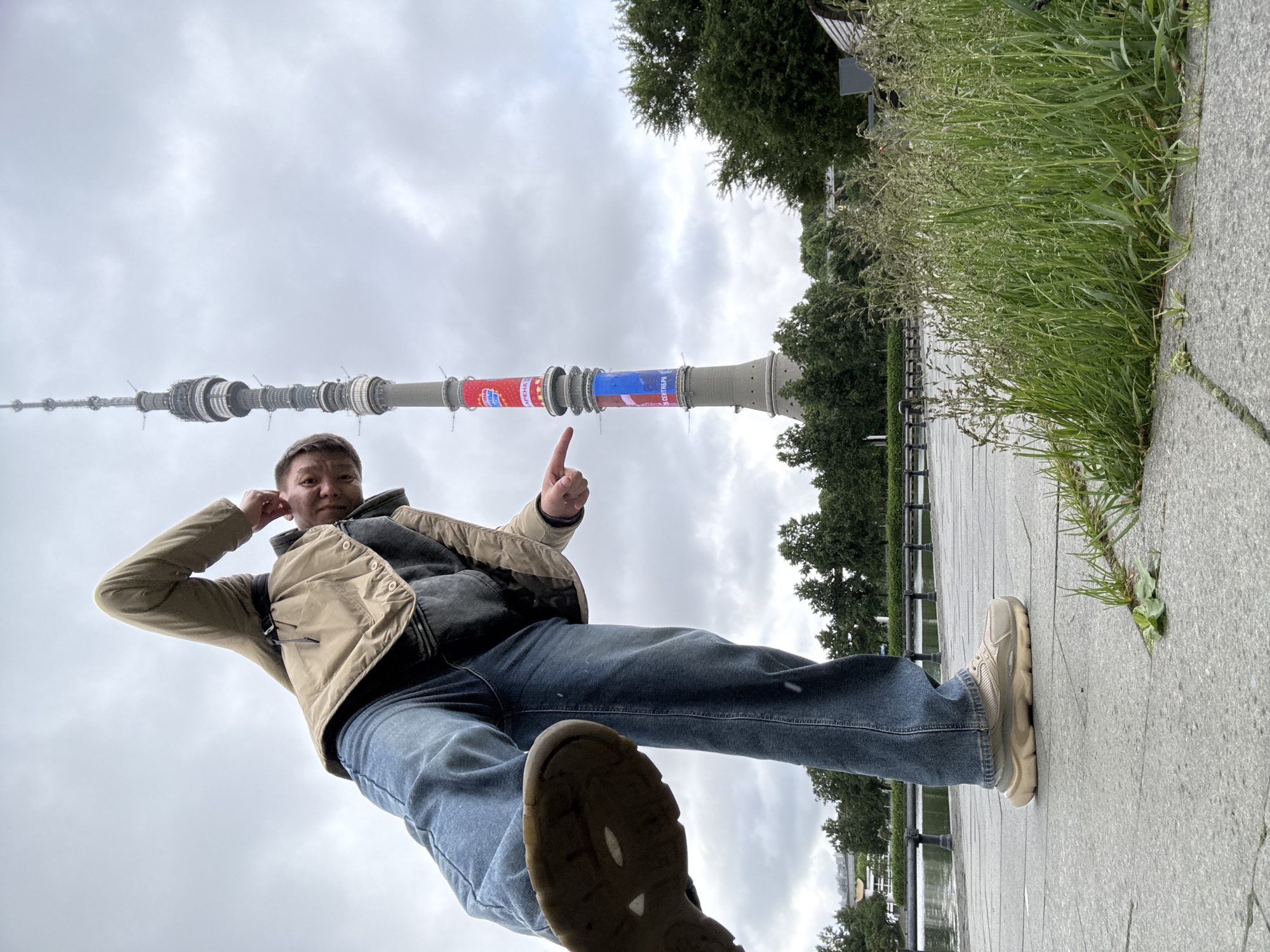A low-angle perspective captures a man standing on a paved surface, with the Ostankino Tower in the background, suggesting a location potentially in Moscow, Russia, rather than Khimki specifically. The sky is overcast, and vegetation, including grass and trees, are present. The man is pointing at the tower with his right hand and has his left hand near his ear. He wears jeans, a vest, and sneakers. The perspective makes him appear to be towering over the landscape.