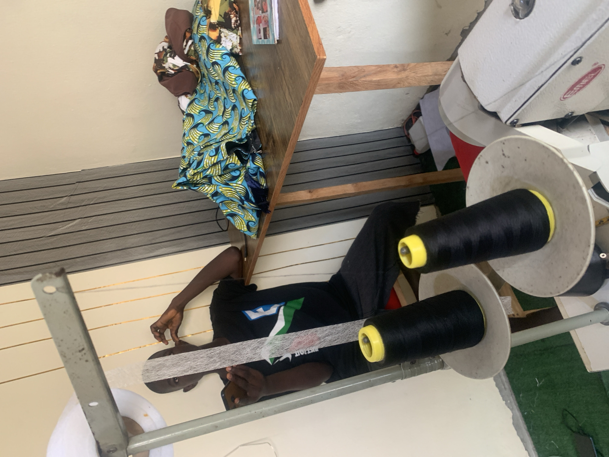 The image depicts a young man sitting in what appears to be a tailor's workshop in Jauro Yinu, Nigeria. He is wearing a black t-shirt and dark pants. In his left hand, he holds a mobile phone, and his right hand is near his face. In front of him, a roll of white material is being fed from a metal stand. Two large spools of black thread with yellow tops are prominently placed on stands in the foreground.
To the right of the man, a wooden table is laden with colorful fabrics, primarily a blue and yellow patterned textile. A book or magazine is also visible on the table. Behind the table, a wall with vertical grey and white stripes serves as a backdrop. A white sewing machine is partially visible in the bottom right corner of the image. The floor appears to be covered with a green mat in some areas. The overall setting suggests a functional workspace dedicated to tailoring.