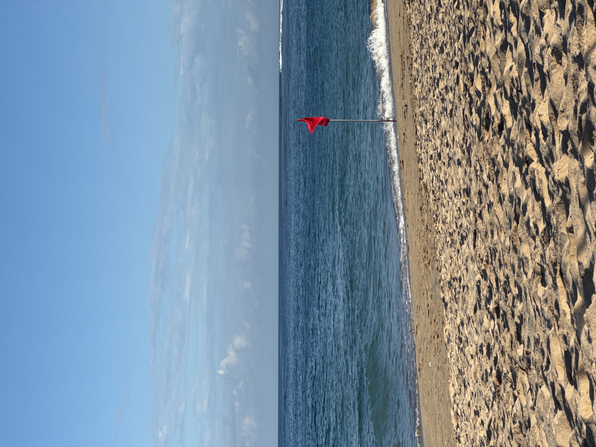 The image depicts a beach scene, with a clear blue sky and scattered clouds visible on the left side. To the right is the ocean, with gentle waves lapping onto the shore. A red flag is planted near the shoreline, likely a warning symbol. The rightmost portion of the image showcases the sandy beach, with uneven textures and shadows. The location is likely in Kuta Selatan, Indonesia, as evidenced by the tropical beach environment. There are no people visible in the image.