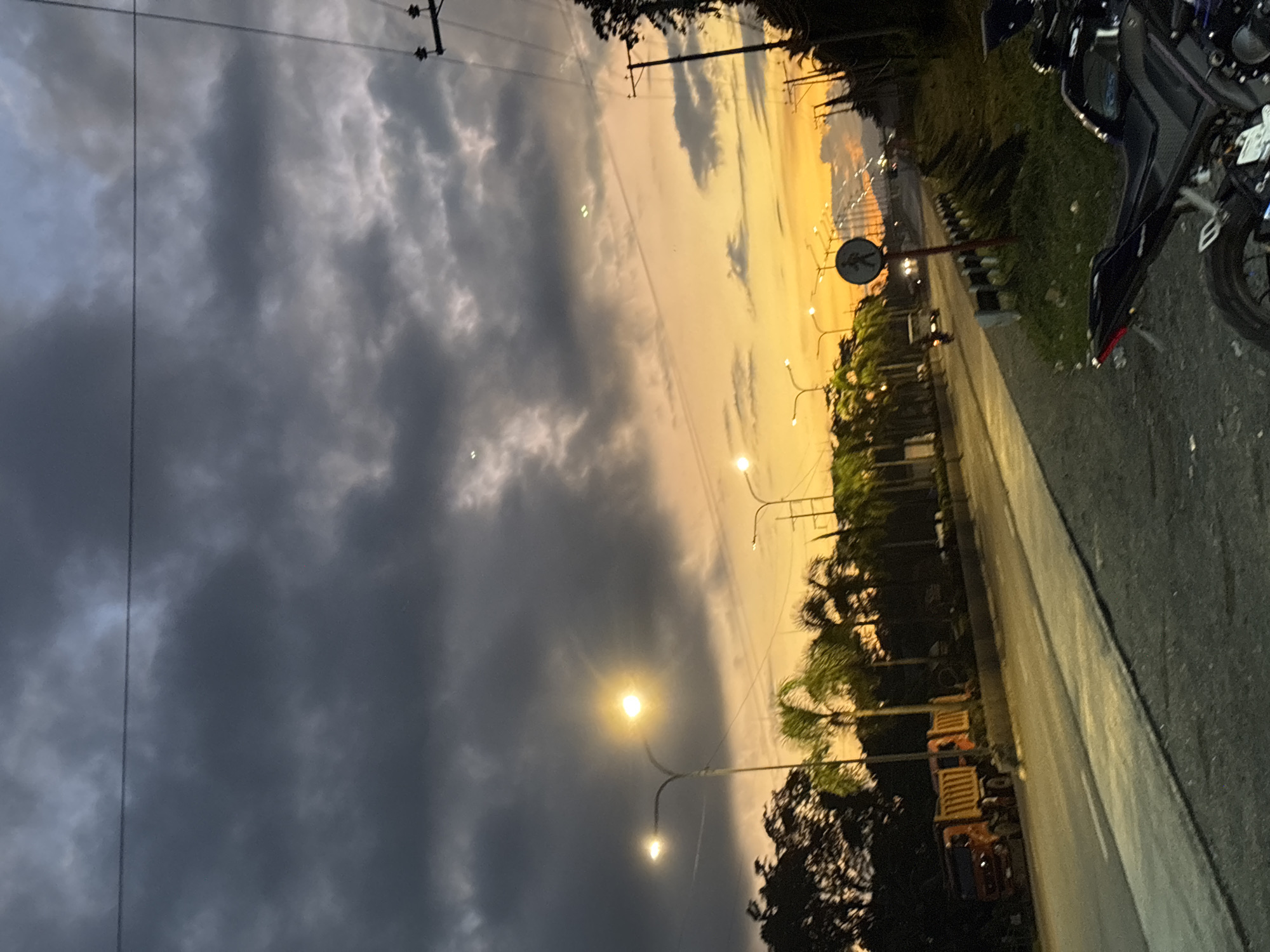 The image depicts a roadside scene at dusk in Carmen, Philippines. The sky is filled with dramatic clouds, ranging from dark gray to vibrant orange and yellow hues, indicating either an impending storm or a sunset. Several streetlights are illuminated along the road, casting a warm glow. In the foreground, a black motorcycle is parked on the side of the road. The road itself is paved and appears to be a highway or main thoroughfare, with a white line marking the lanes. To the left of the road, a row of palm trees and other foliage is visible, silhouetted against the colorful sky. Several vehicles, including a red truck and a white van, are present on the road, suggesting moderate traffic. A pedestrian crossing sign with a silhouette of a person walking is also visible. In the background, more palm trees and vegetation line the side of the road, and distant mountains or hills are faintly visible. Utility poles and power lines are present on the right side of the image, typical of Philippine infrastructure. The overall atmosphere is serene and slightly moody due to the dramatic lighting conditions.
