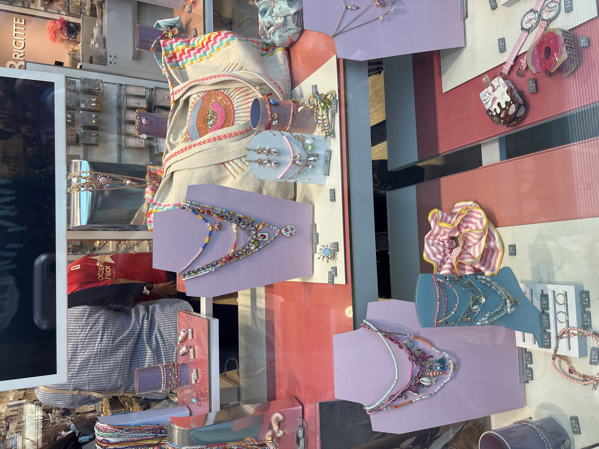 This image shows a display window of a jewelry store, possibly named Brigitte, located in Orihuela, Spain. The display features a variety of necklaces, bracelets, earrings, and a large tote bag adorned with an eye motif. The items are arranged on purple and white stands and on a white surface. In the background, a person wearing a blue and white striped shirt and another person wearing a red shirt are partially visible. The display also includes price tags for the jewelry. The overall environment appears to be a well-lit store interior.