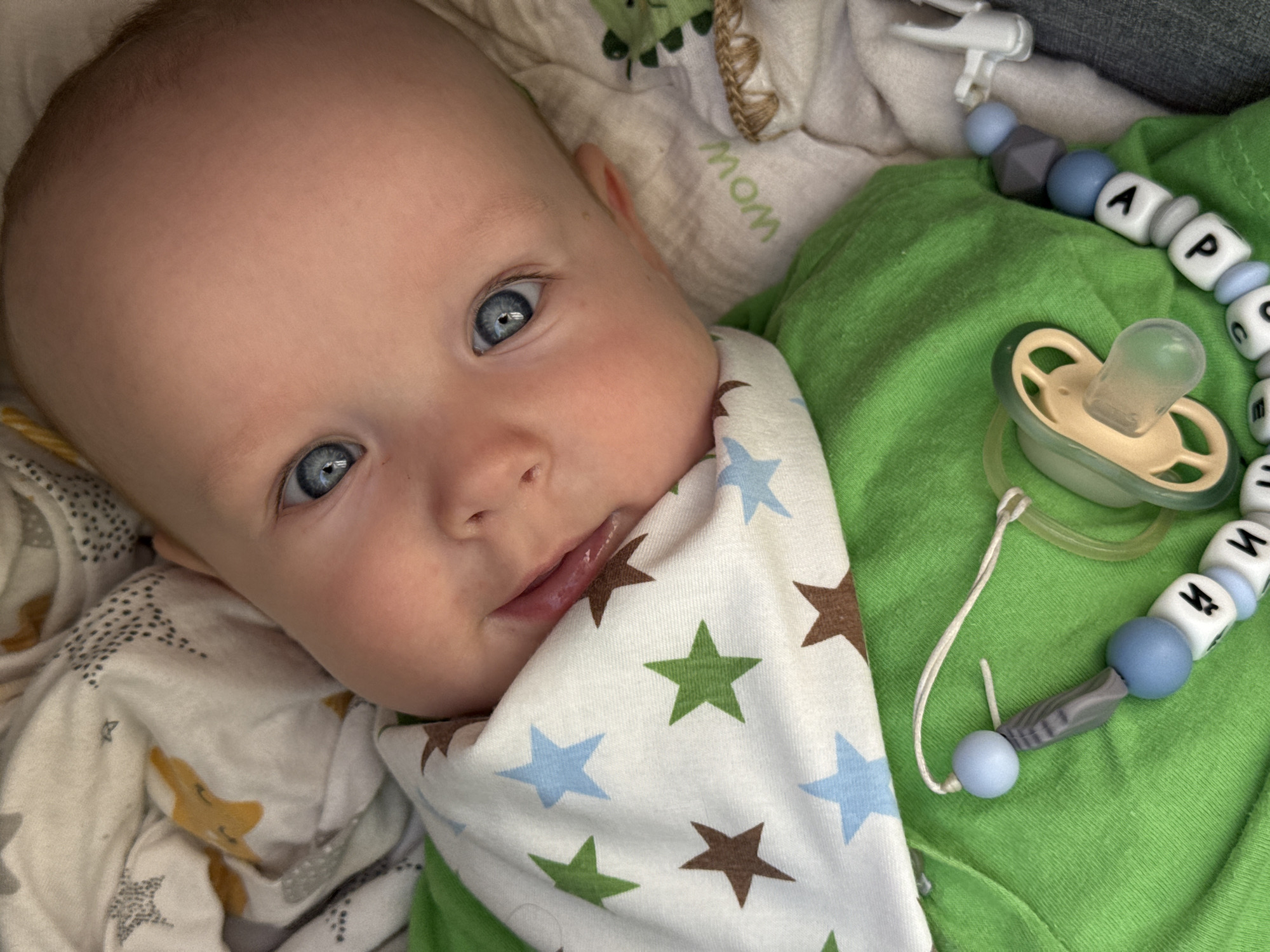 This image depicts an infant, approximately 4-8 months of age, resting in an indoor setting within the city of Moscow, Russia. The infant has fair skin, light blue eyes, and minimal light-colored hair, and is positioned looking directly at the viewer with a slight, open-mouthed expression revealing the tongue.
The infant is dressed in a bright green short-sleeved shirt and a white bib patterned with alternating green, light blue, and brown five-pointed stars. A light green pacifier, featuring a clear silicone nipple and a green-grey plastic ring, is attached to a custom beaded chain. This chain comprises light blue and grey spherical and polygonal beads, interspersed with white cubic beads bearing individual letters, including "И," "Н," "К," "Т," "Е," "М," "А," "Р," and "С." The chain's clip mechanism is partially visible on the far right, securing it to an unseen anchor.
The infant is supported by soft bedding. To the upper left, a white fabric with patterns of grey stars, yellow animal figures (resembling foxes), and black speckled dots is visible. To the right, a cream-colored fabric with green embroidered text "WOW" and a distinct brown braided detail is present. The overall environment appears to be a well-lit, comfortable indoor space.