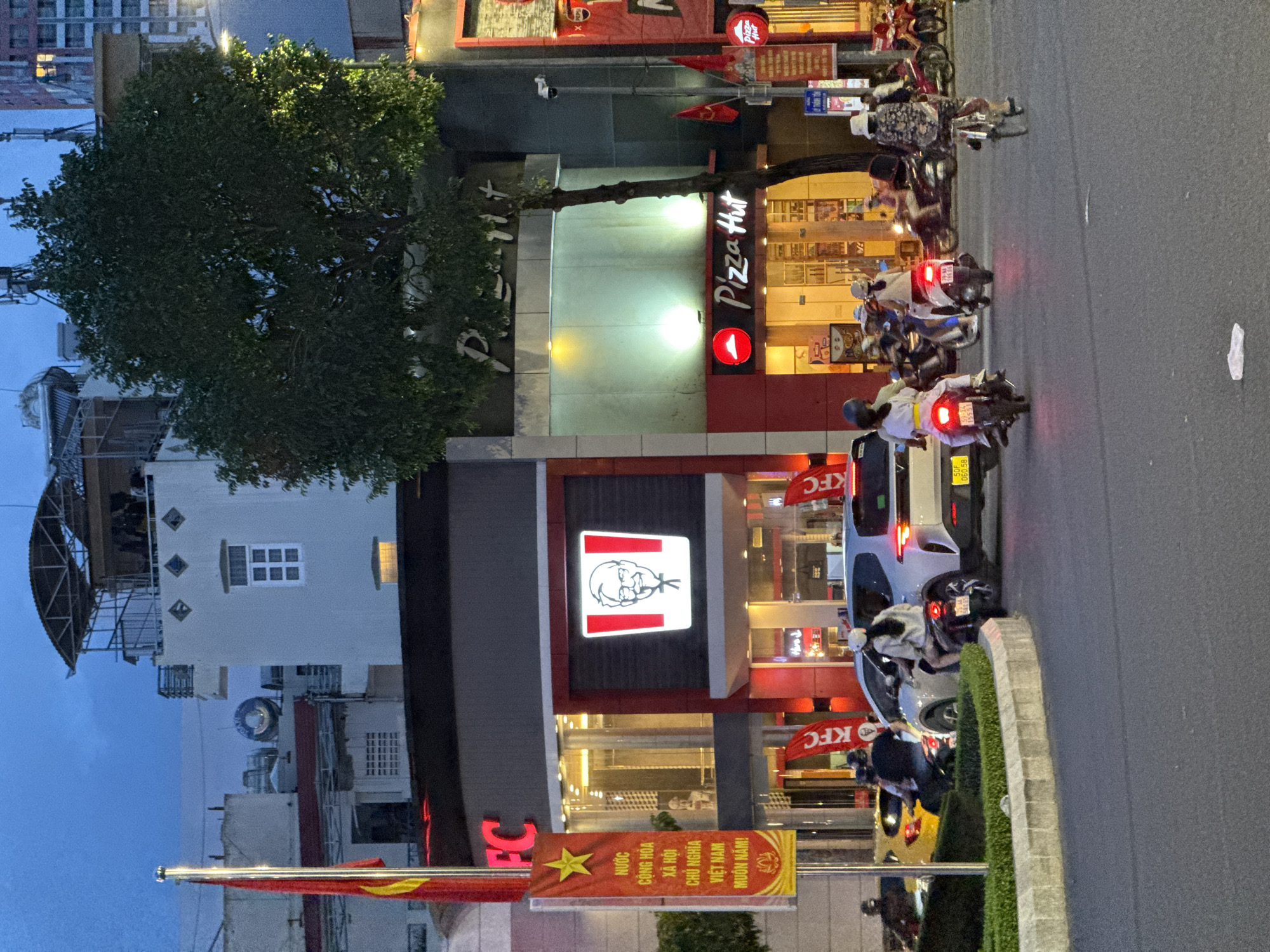 The image shows a street scene at dusk in Nha Trang, Vietnam, featuring two prominent fast-food establishments: KFC and Pizza Hut. A large KFC sign with the iconic Colonel Sanders logo is clearly visible, as is the Pizza Hut logo. Several people are on motorcycles navigating the street. One motorcycle has a passenger wearing a white helmet and yellow sash, while another has a passenger wearing a patterned dress and a hat. In the background, buildings and trees are illuminated by streetlights. A Vietnamese flag is also present, flying from a pole on the left side of the frame. The street appears to be a multi-lane road with a concrete divider adorned with greenery. The sky is a dusky blue, indicating the transition from daylight to night.