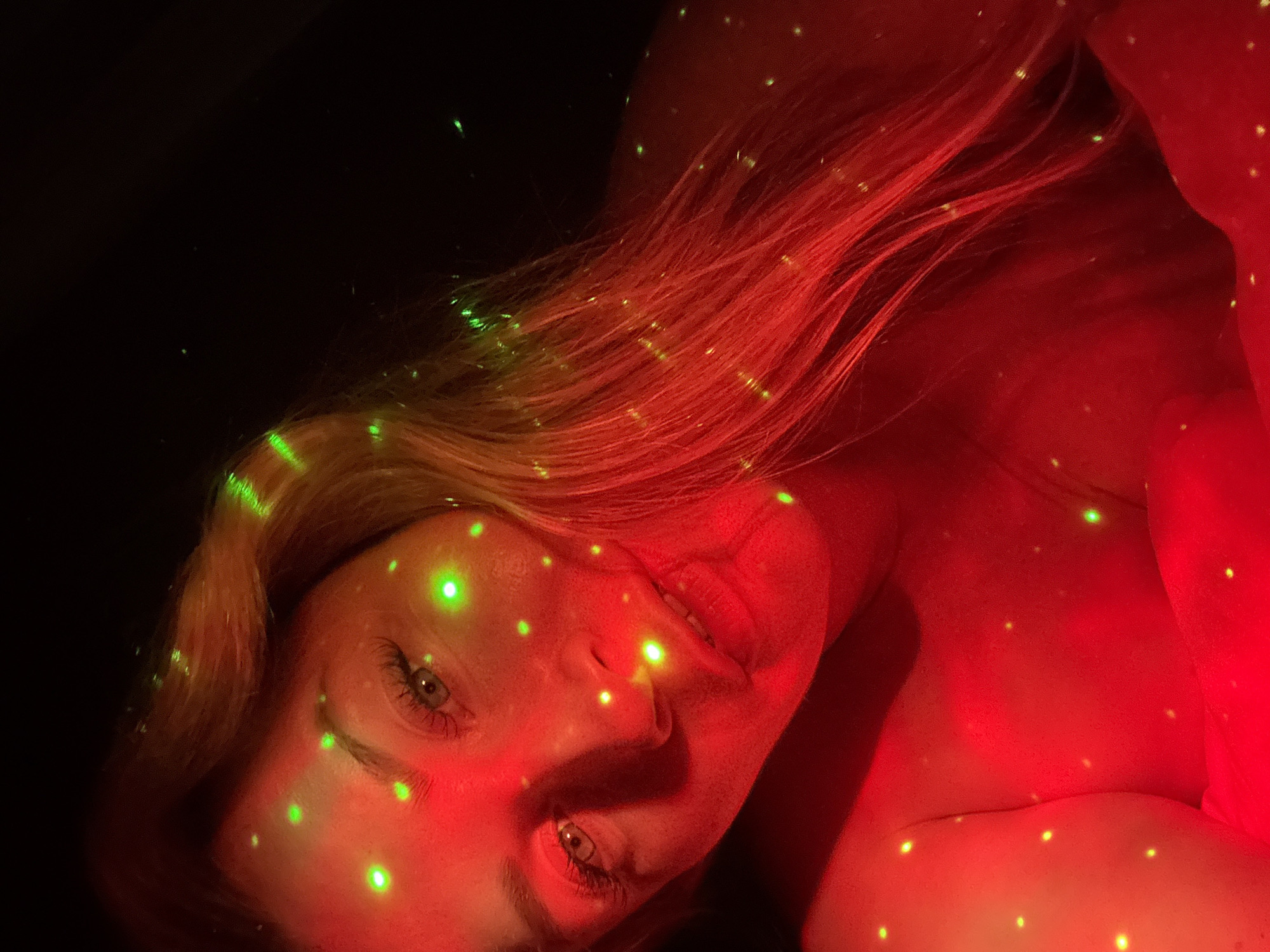 The image is a close-up portrait of a person's face, illuminated by red and green lights, creating a starry effect. The person appears to be female, with blonde hair that is partially visible. The lighting obscures facial features in some areas, but the eyes and lips are discernible. The background is predominantly black, suggesting a dimly lit environment. There is no discernible connection to the city of Veliky Novgorod, Russia.