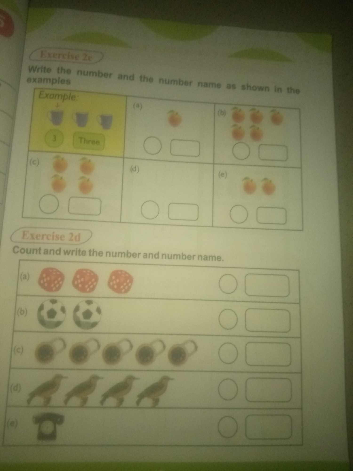 The image displays a page from a workbook with two exercises, "Exercise 2c" and "Exercise 2d". "Exercise 2c" instructs the user to write the number and number name as shown in the examples. The example shows three cups, with the number "3" and the word "Three" written below. The exercise then presents images of fruits in boxes labeled (a), (b), (c), and (e), each with two empty spaces for the user to fill in the number and name. "Exercise 2d" asks the user to count and write the number and number name. It shows images of dice, soccer balls, padlocks, birds, and a telephone, with empty spaces for the user to write their answers. The location of Jauro Yinu, Nigeria is not depicted or referenced in the image.